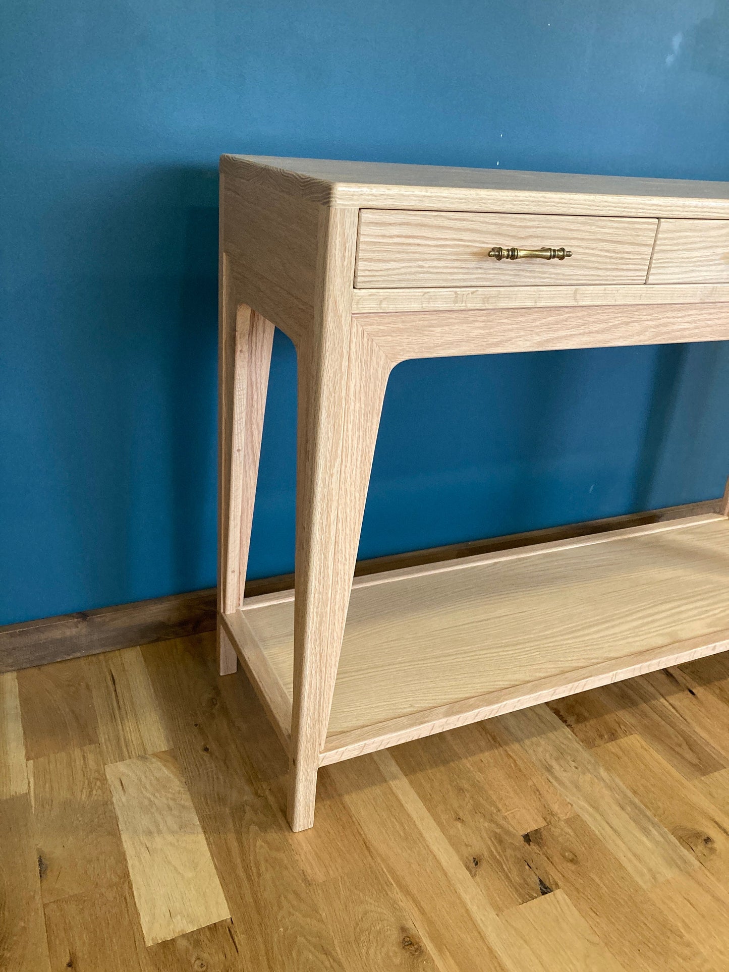 A light wood console table with drawers and a lower shelf. The drawers have brass handles. The table is angled and has a modern design.