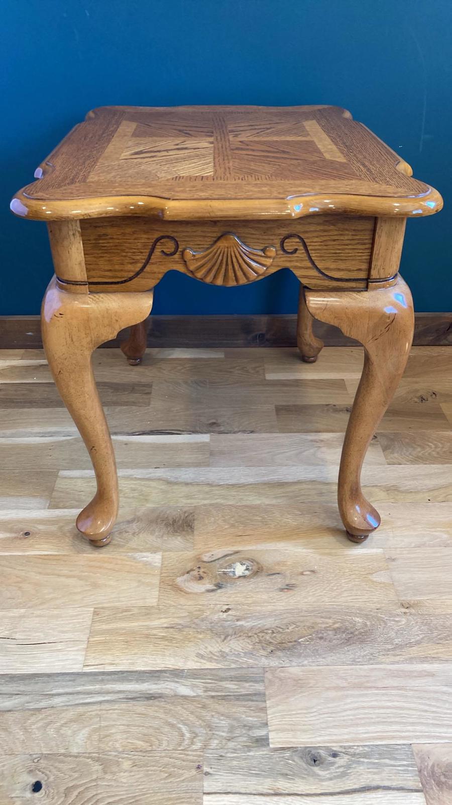 A light brown wooden side table with cabriole legs. The top features an inlaid parquetry design. The front is adorned with shell-shaped carvings. The table is shown on a light wood floor against a teal background.