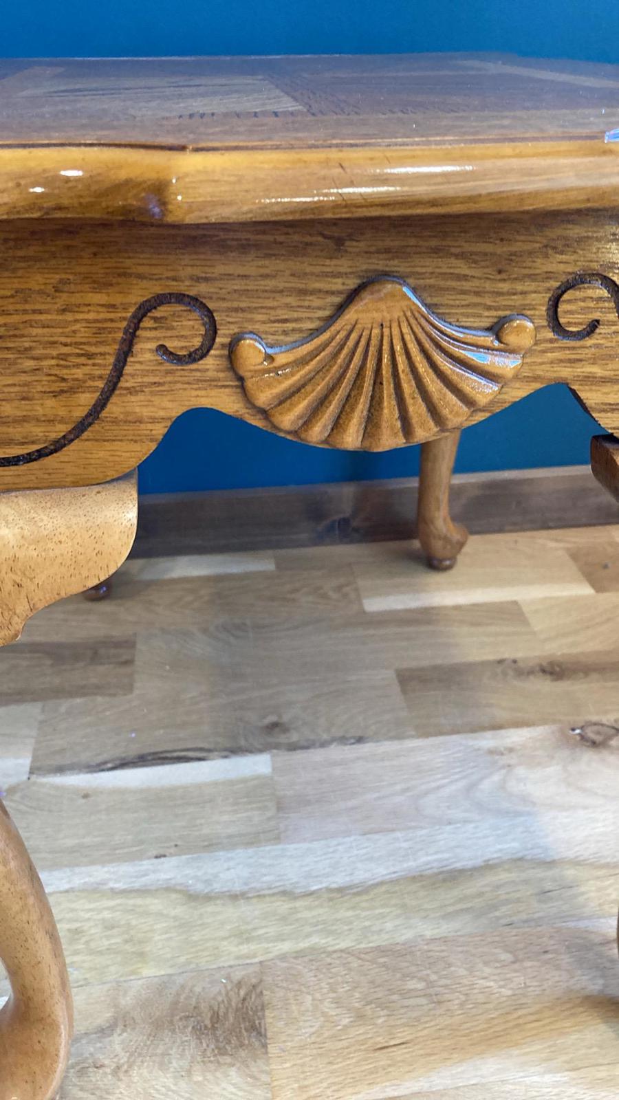Close-up of a wooden coffee table with ornate carvings. The table features a shell-shaped carving at the center and scrollwork details on the apron. The legs are cabriole style, and the top is a rich, medium brown wood. The table sits on a light brown wood floor.