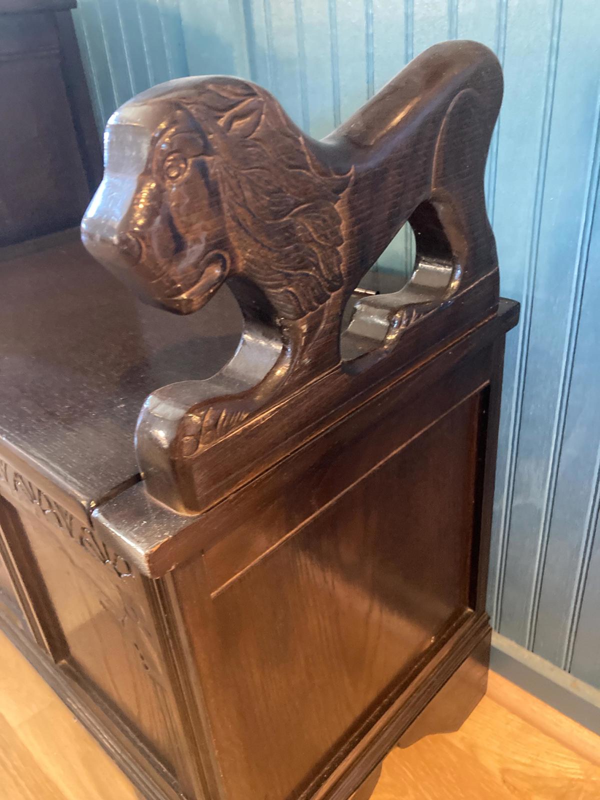 Dark brown wooden storage bench with intricately carved lion head armrests. The bench has a rectangular shape with a hinged top, offering storage space. The lion carvings are detailed, showing mane and paws. The wood grain is visible, and the overall style suggests a vintage or antique aesthetic. The bench is shown against a blue-green wall.