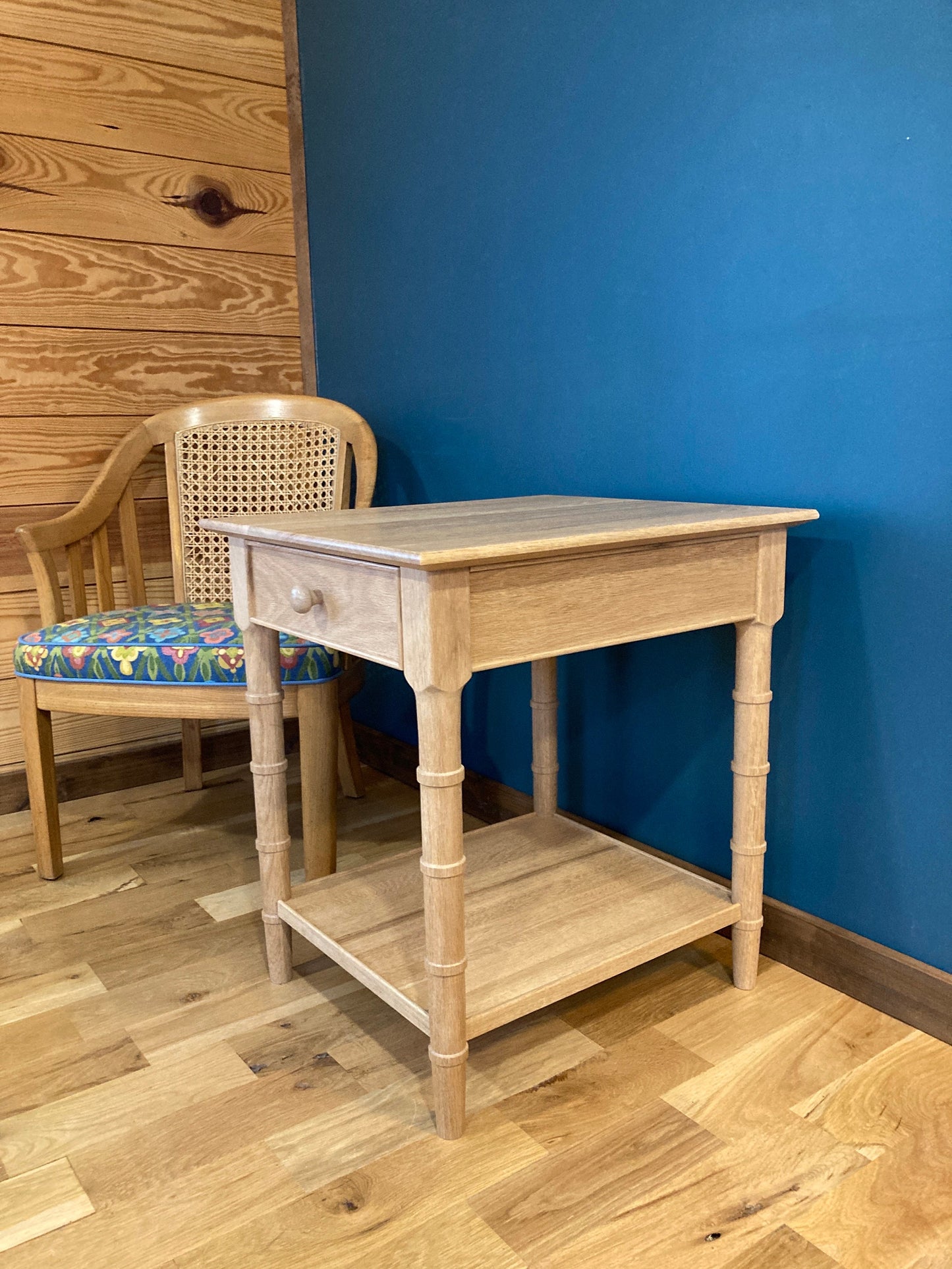A light wood end table with a single drawer and a lower shelf. The table has a simple design with turned legs. The table is in front of a blue wall and a wooden floor.