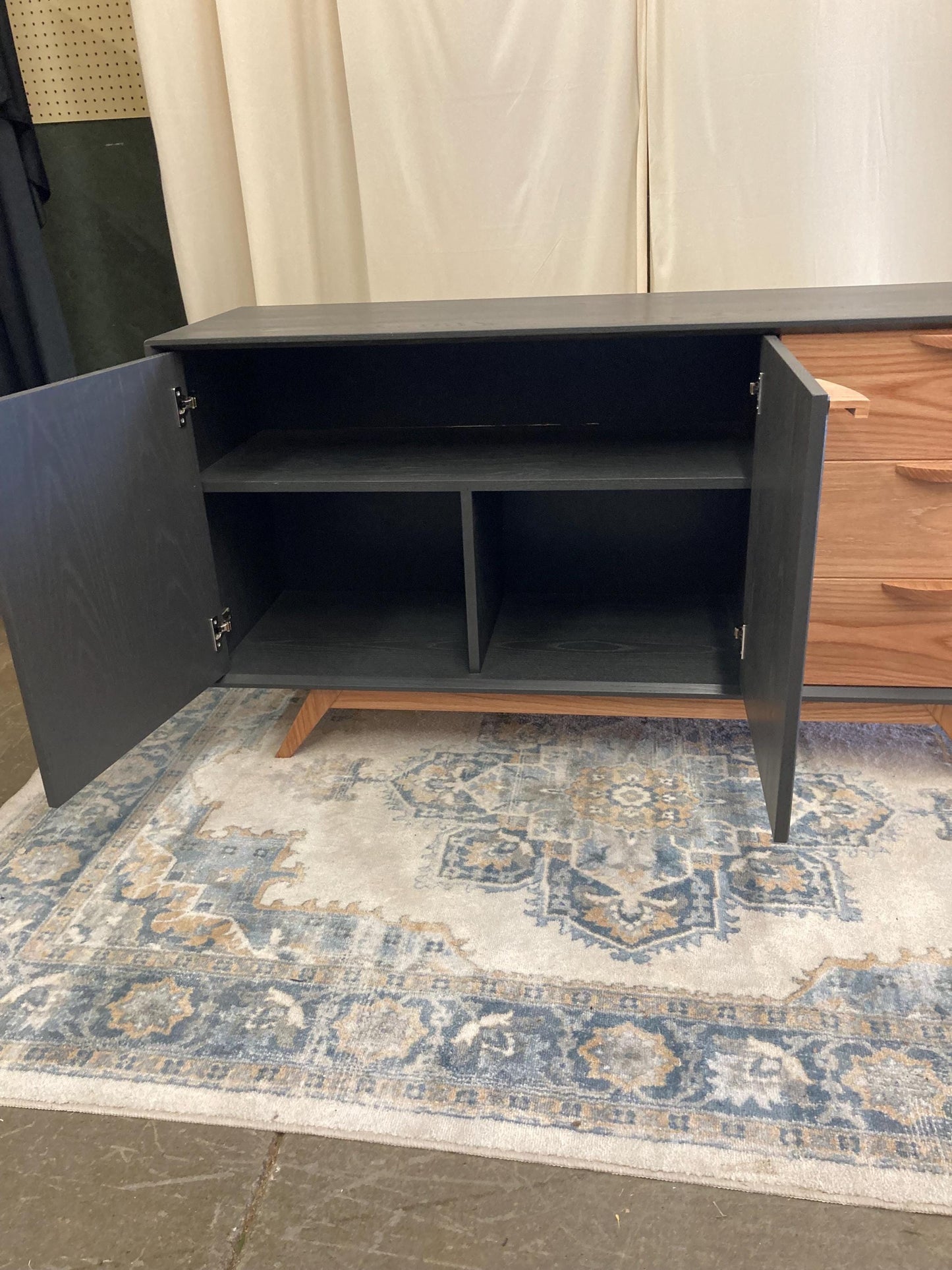 A gray and light wood sideboard with two cabinet doors and three drawers. The cabinet doors are open, revealing adjustable interior shelving. The sideboard features mid-century modern style legs. The piece is displayed on a blue and beige patterned rug.