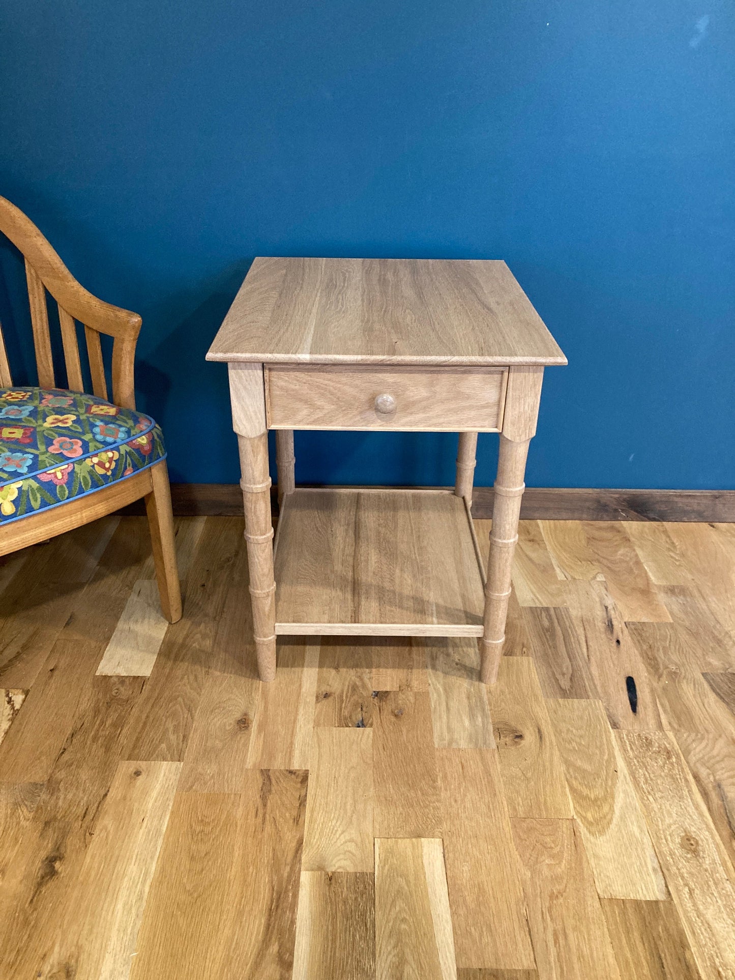 A light wood end table with a single drawer and a lower shelf. The table has turned faux bamboo legs and is sitting on a wood floor.