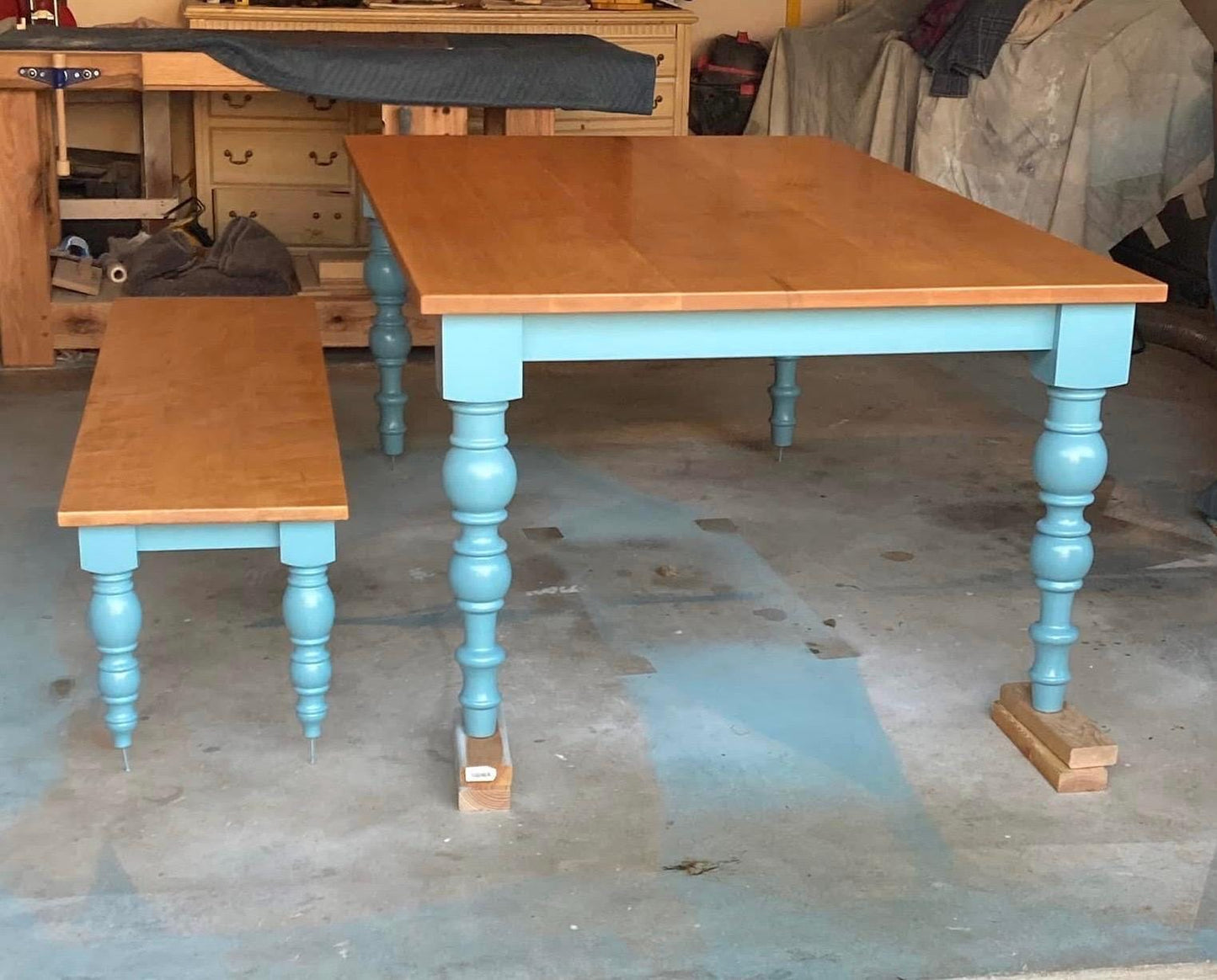 A light blue wooden table with a natural wood top and turned legs. A matching light blue wooden bench with a natural wood top and turned legs is in front of the table.