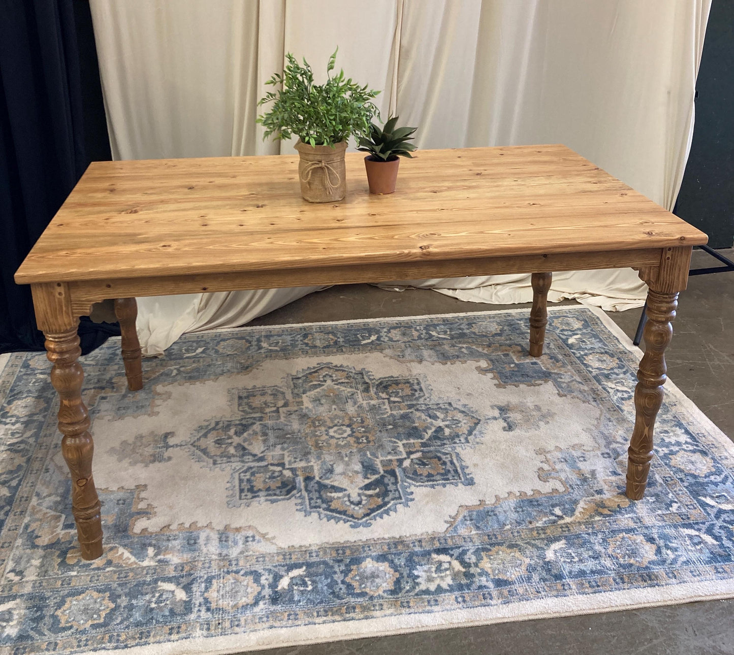 A rustic wooden table with a light brown finish. The table has four turned legs and a rectangular top. The table is sitting on a rug with a blue and beige pattern.