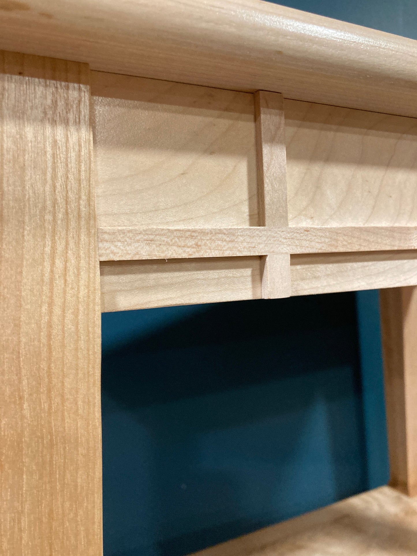 Close-up view of a light beige wooden table top with a minimalist, geometric design. The wood grain is visible, and the piece features clean lines and a simple, rectangular frame with a cross-shaped detail. The shelf's top edge has a subtle rounded profile. The background is a dark teal or blue-green.