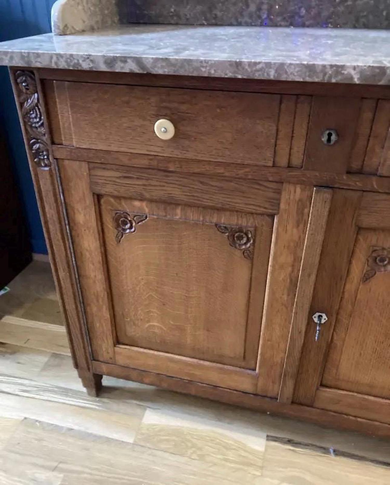 A wooden cabinet with a marble top. The cabinet has two doors and two drawers. The doors are closed and have carved floral designs. The drawer is closed and has a small, round knob. The cabinet is a light brown color and the marble top is a light brown color.