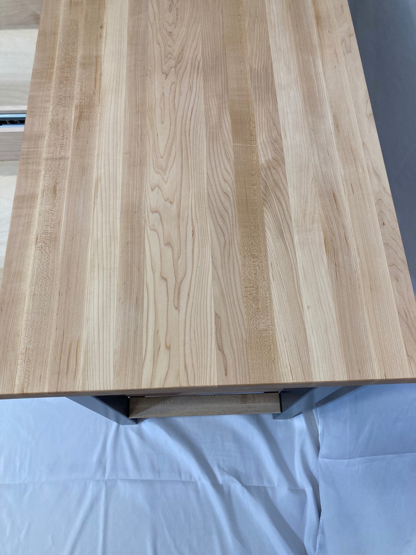 Overhead close-up view of a light-colored wood butcher block kitchen island countertop. The wood grain is clearly visible, running vertically along the surface. The countertop is rectangular and is made of solid wood planks. The island has a simple, minimalist design with a blue base visible underneath. The wood has a natural, light beige and tan color with subtle variations in tone. The overall impression is one of clean, modern design and high-quality materials.