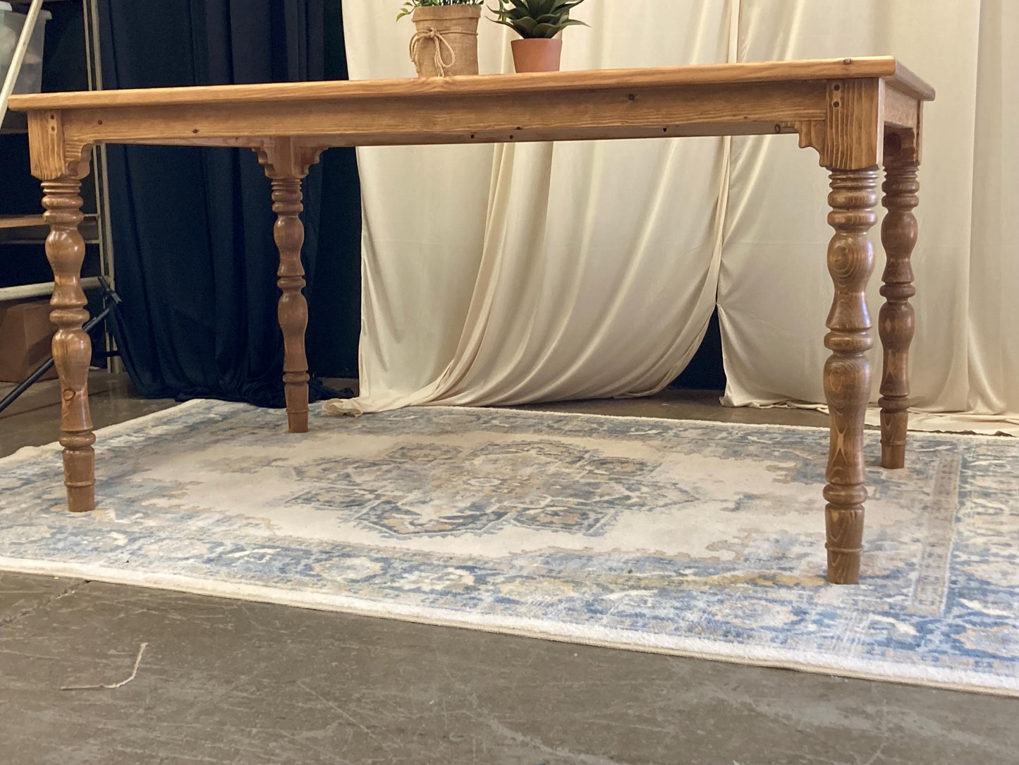 A rustic wooden table with a light brown finish. The table has four turned legs and a rectangular top. The table is sitting on a light blue and cream patterned rug.