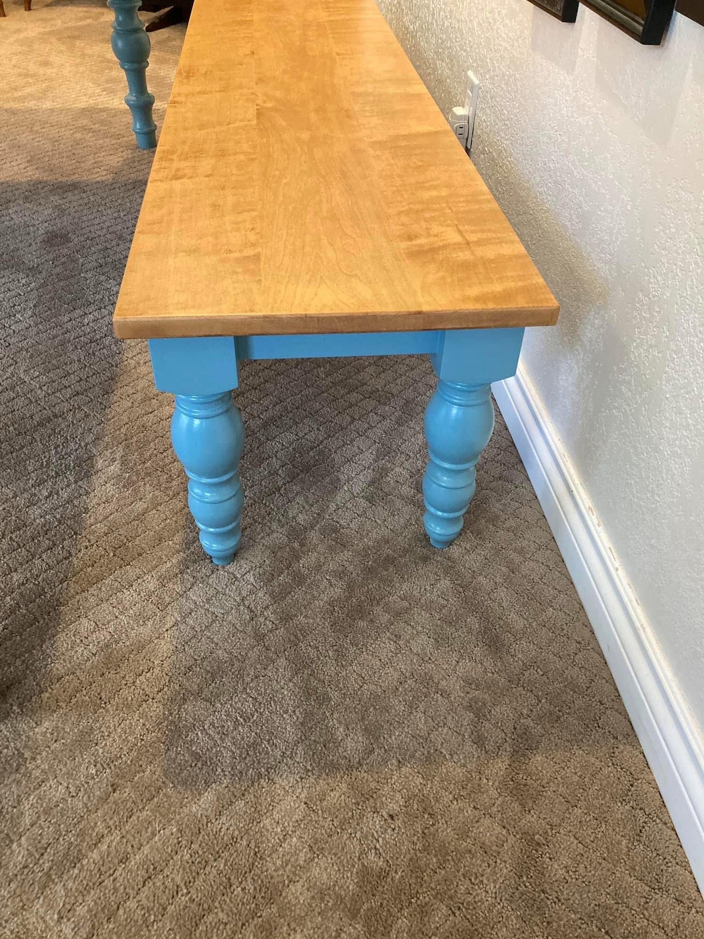 A wooden bench with a light wood top and blue painted legs. The legs have a turned design and are on a brown carpet.