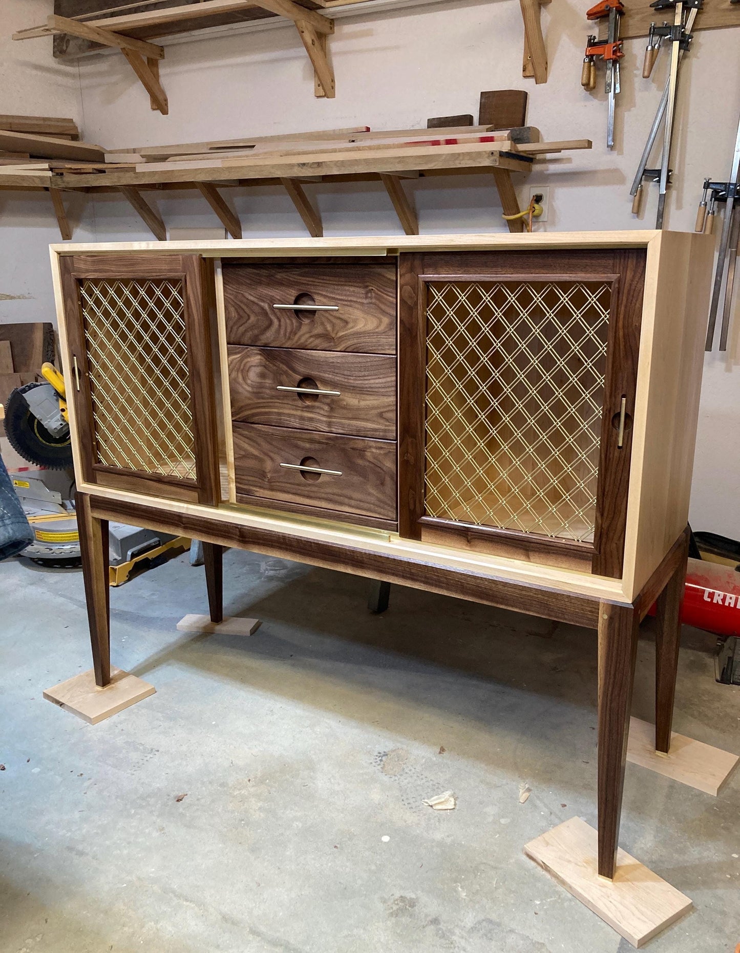 A wooden cabinet with two sliding doors and three drawers. The doors are made of wood with a gold wire mesh insert. The cabinet is standing on four legs and has a light wood top. The cabinet is in a workshop setting.