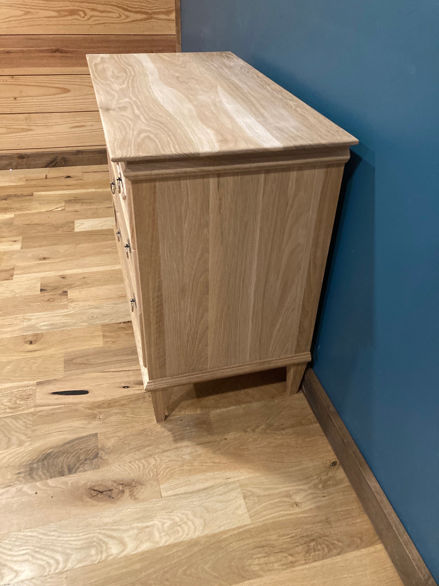 A light oak wood dresser with three drawers and a top surface. The dresser is on a light wood floor and is positioned against a blue wall.