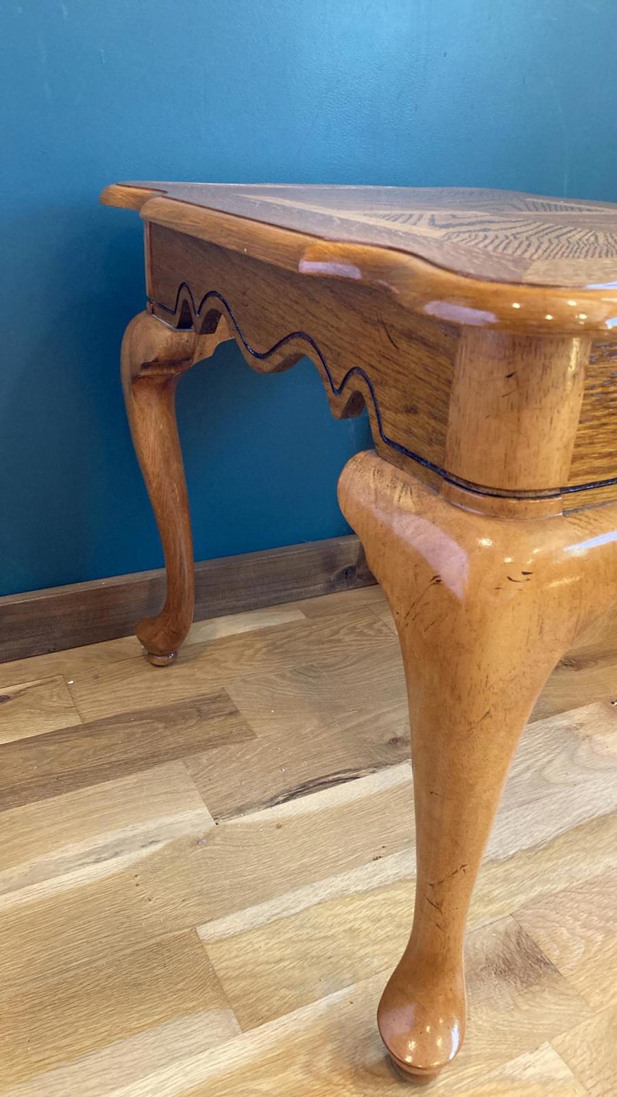A light brown wooden side table with cabriole legs and a scalloped apron. The top is inlaid with a darker wood. The table is shown against a teal wall and light brown wood floor. The table is a classic style and would be suitable for a living room or bedroom.