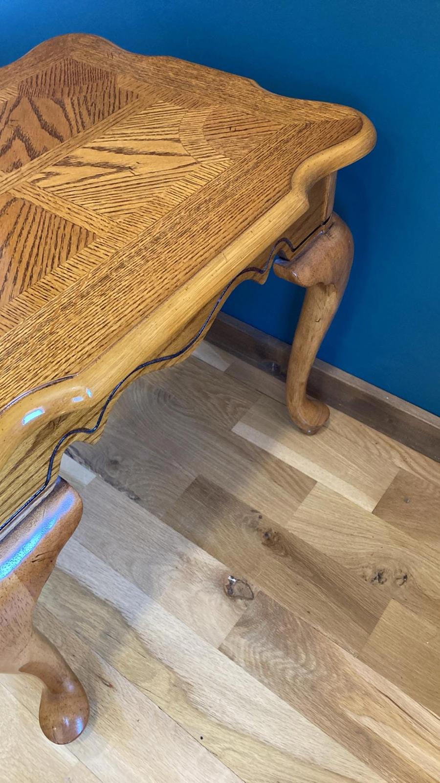 A charming, light brown wooden side table with cabriole legs and a uniquely shaped top. The table's top features an intricate, inlaid wood grain pattern. The edges of the table top are curved, adding to its elegant design. The table is shown against a teal wall and light brown wood flooring.