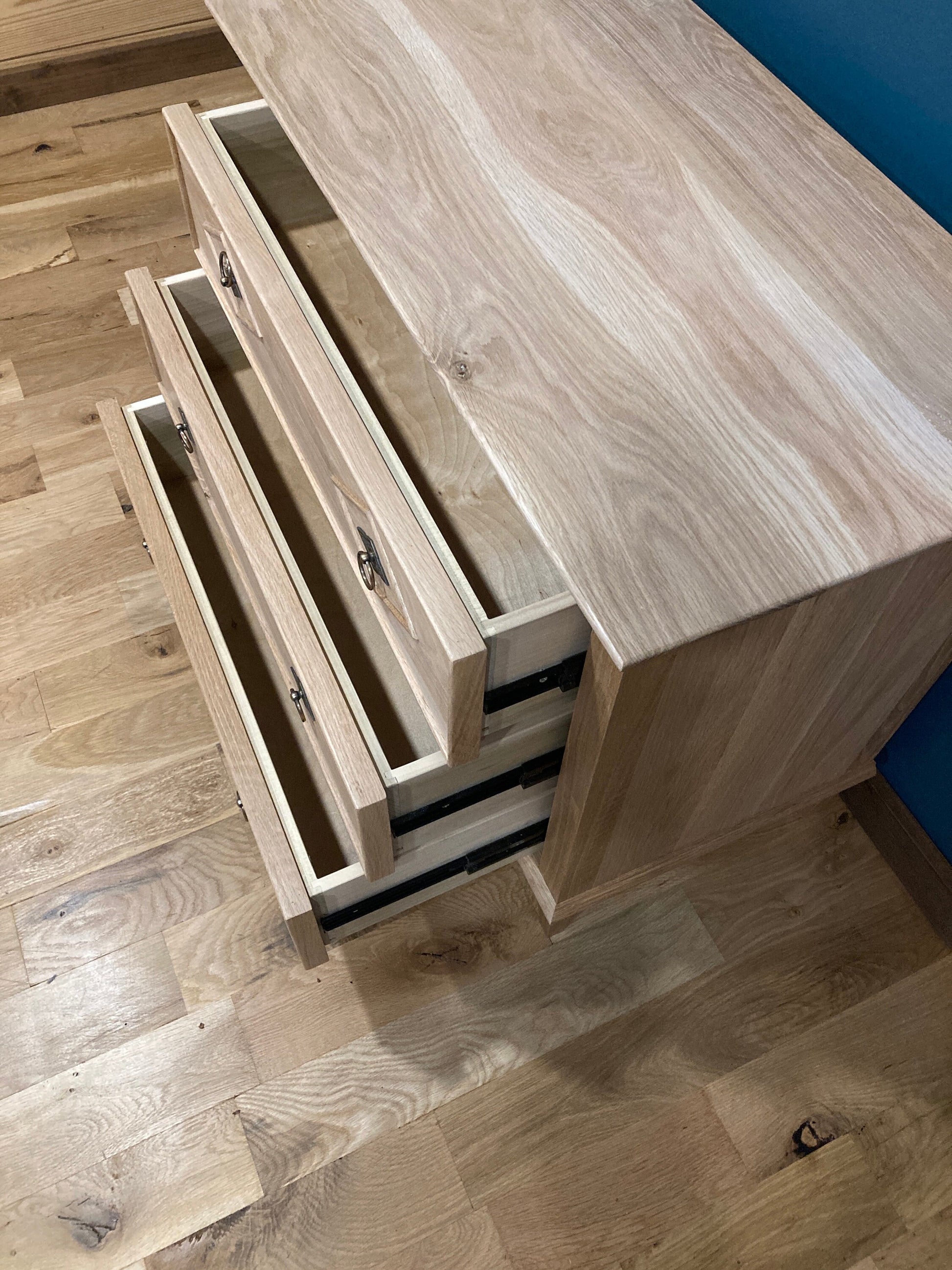 A wooden nightstand with three drawers that are open. The drawers are made of light wood with a natural grain pattern. The dresser is sitting on a light wood floor.