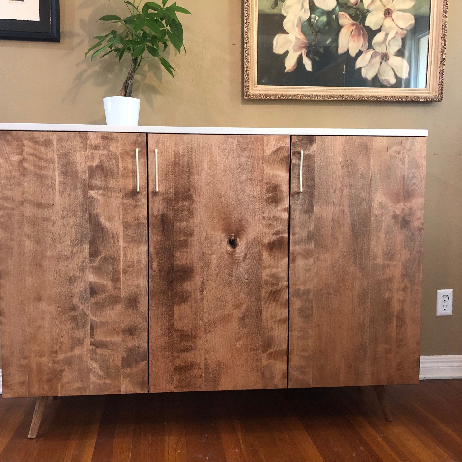 A three-door cabinet with a white quartz stone top and wooden doors. The cabinet has a light brown wood grain finish and silver door handles. The cabinet is sitting on a wooden floor.