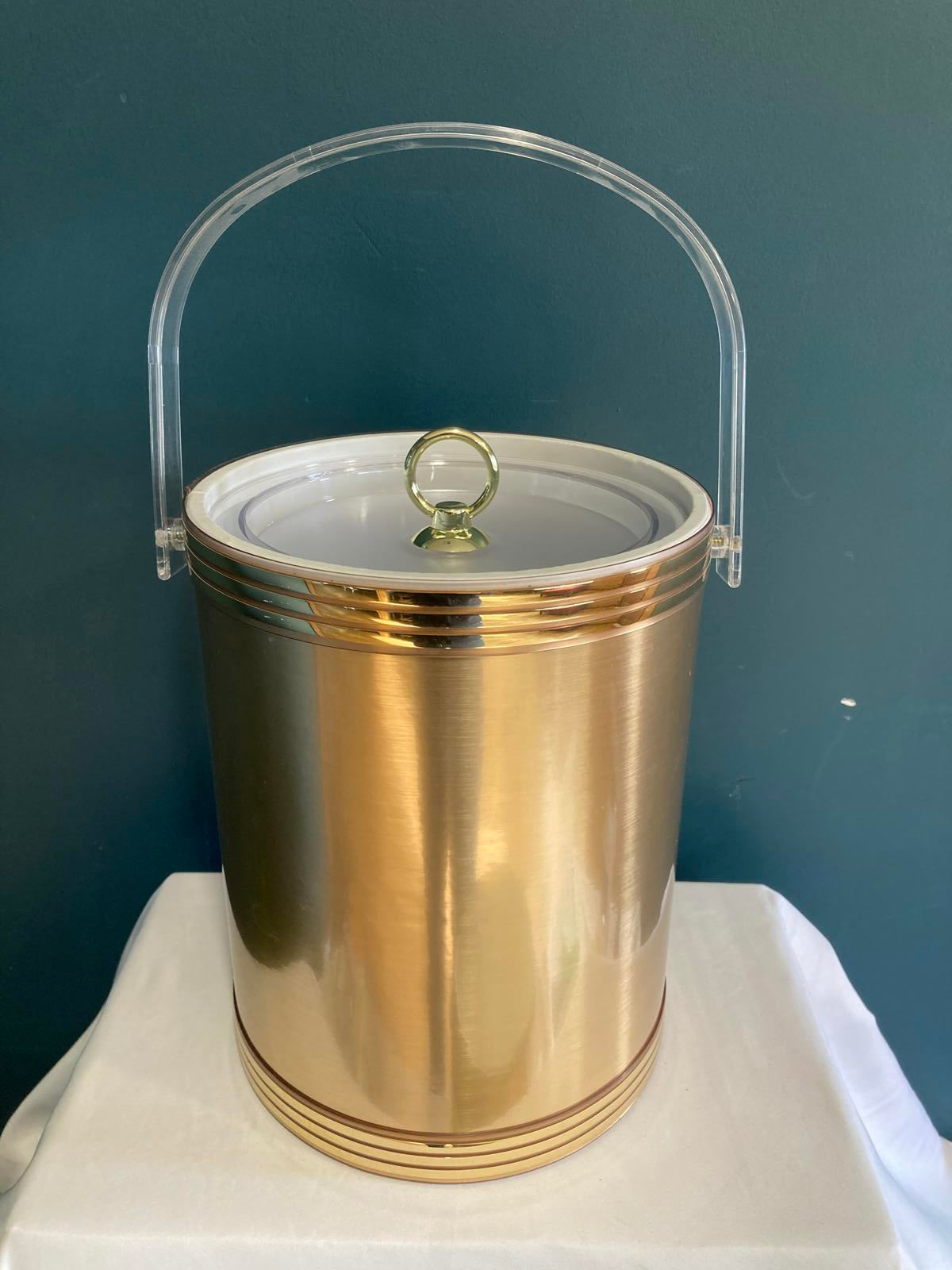 Gold cylindrical midcentury ice bucket with a clear cover on a blue background