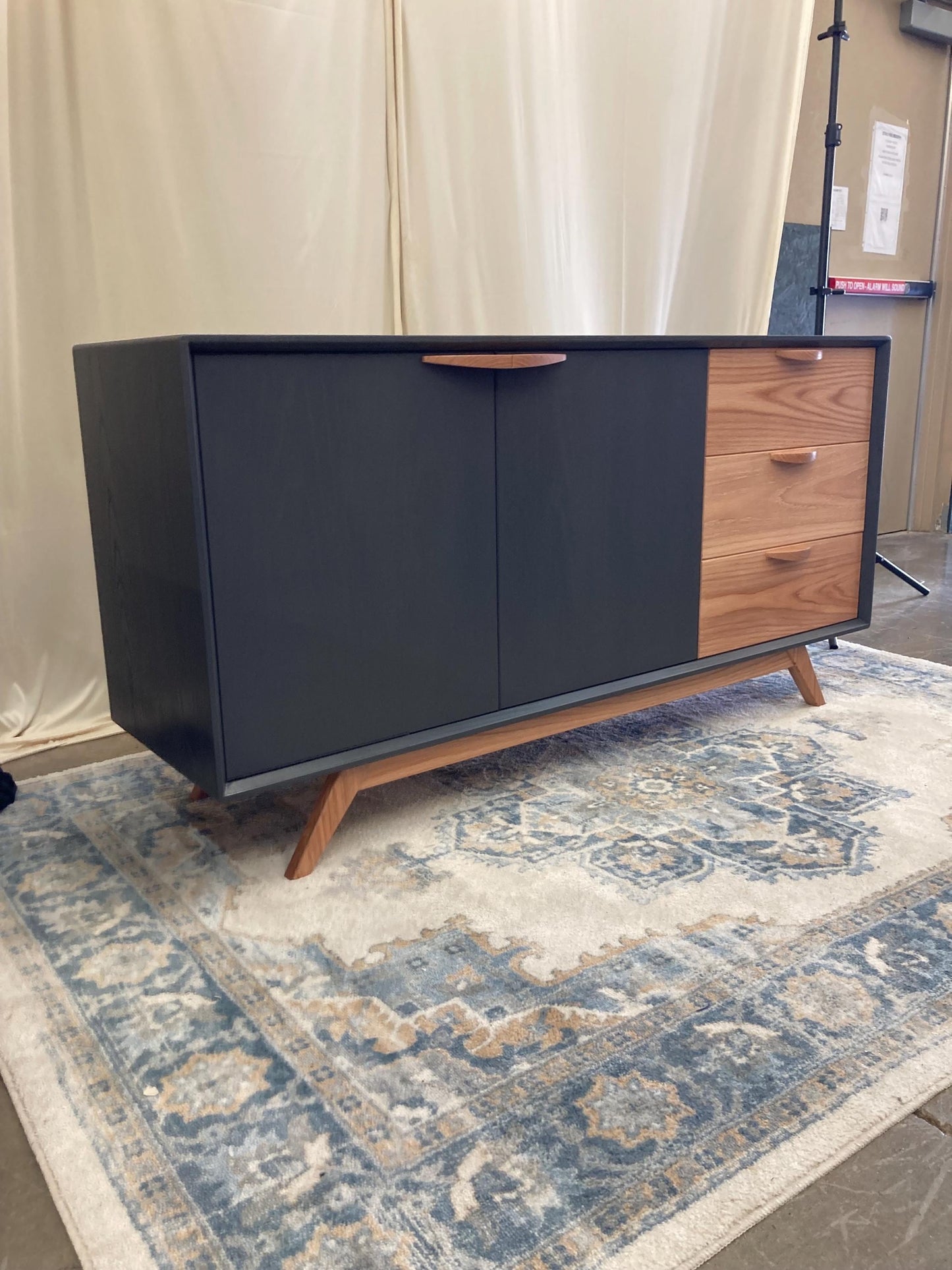 A mid-century modern sideboard with a two-tone design. The left side is dark gray, while the right side features three drawers in light brown wood.  The cabinet features sleek, tapered legs and minimalist wood handles. The sideboard is displayed on a patterned rug.