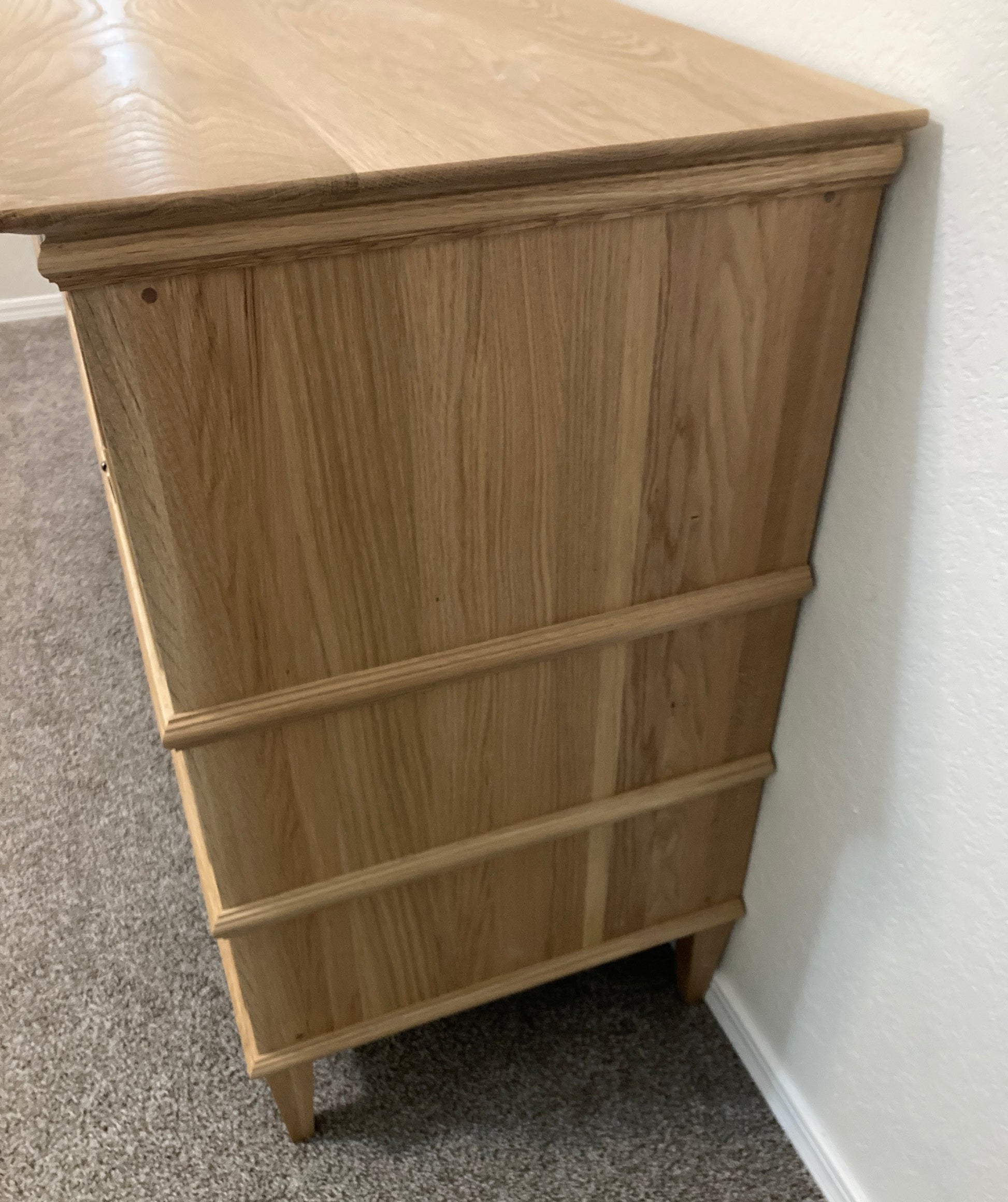 Side view of a light wood dresser with eight drawers. The dresser has a simple design with a slightly raised top edge and a decorative trim around the drawers. The dresser is standing on a light gray carpet.