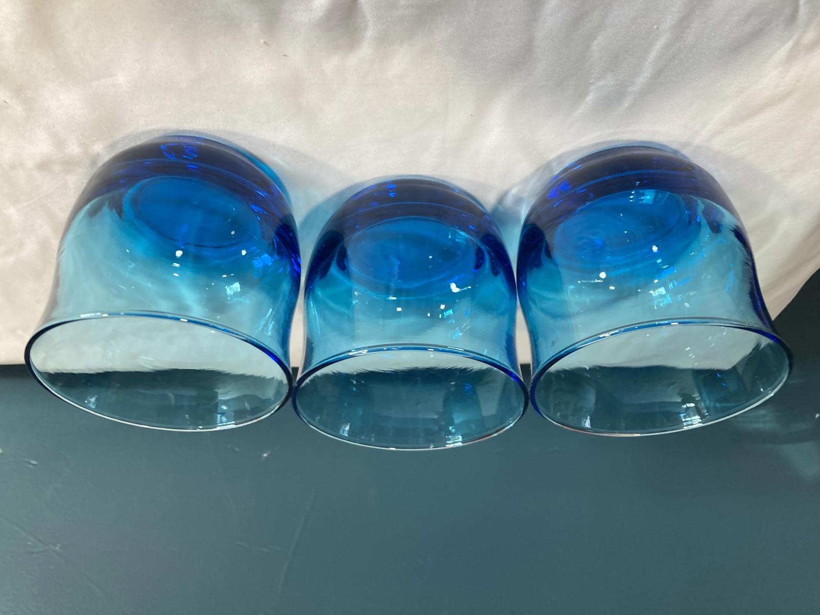Three blue glass tumblers on a surface with a white fabric background