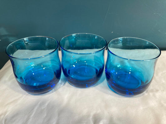 Three blue glass tumblers on a surface with a white fabric background