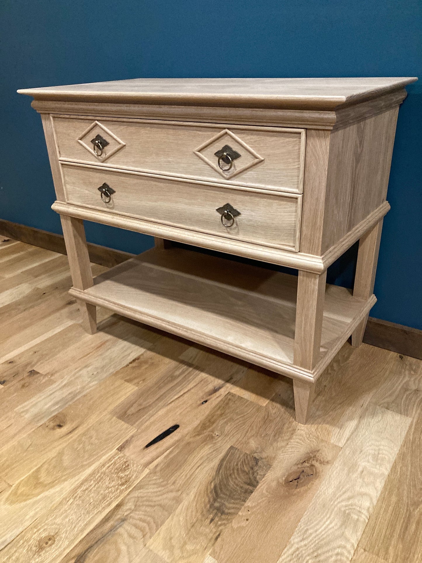 Custom hardwood nightstand with two drawers featuring diamond-patterned accents and vintage-style metal pulls, plus an open lower shelf, shown against a teal wall on hardwood flooring.