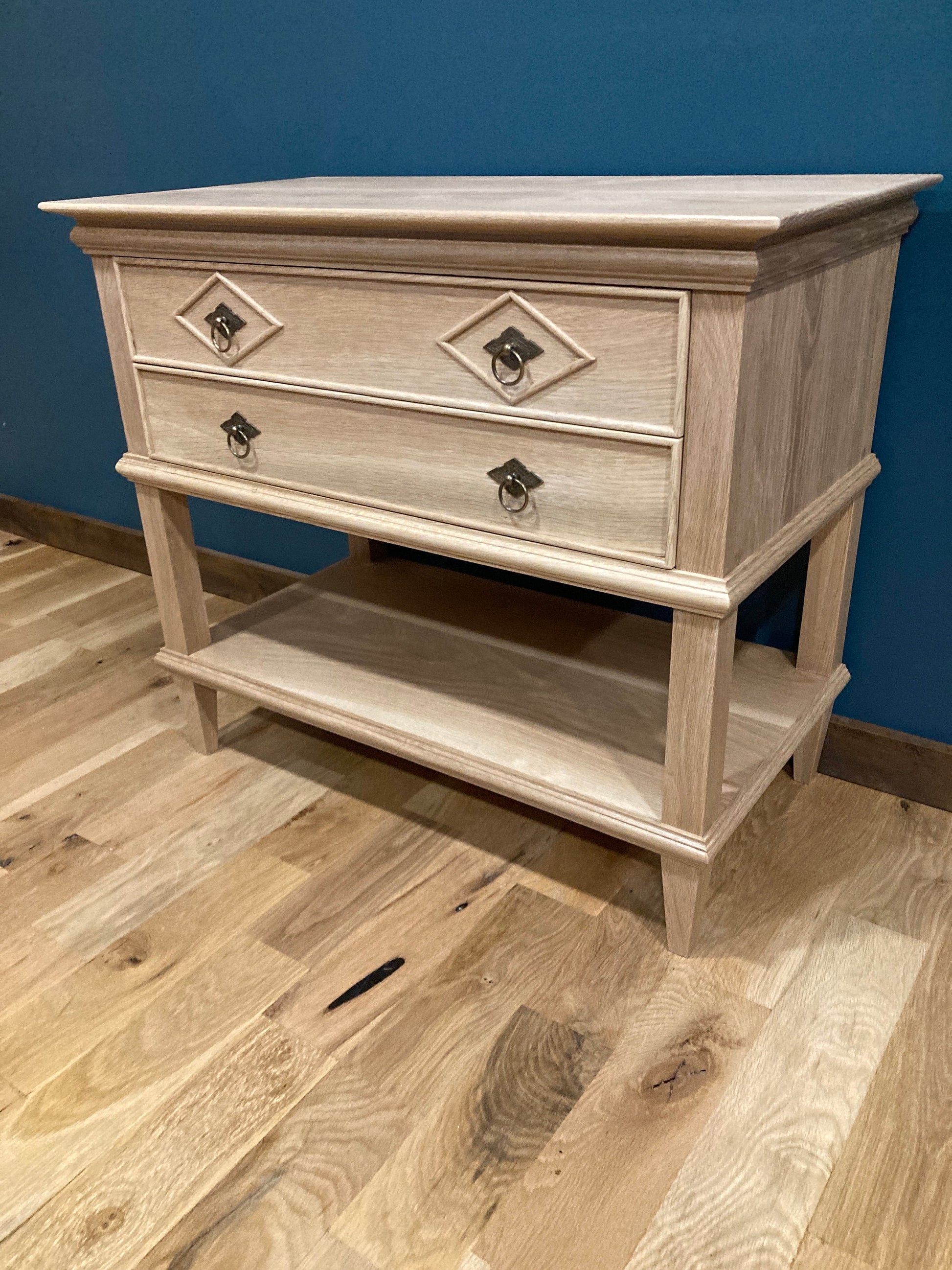 Custom hardwood nightstand with two drawers featuring diamond-patterned accents and vintage-style metal pulls, plus an open lower shelf, shown against a teal wall on hardwood flooring.