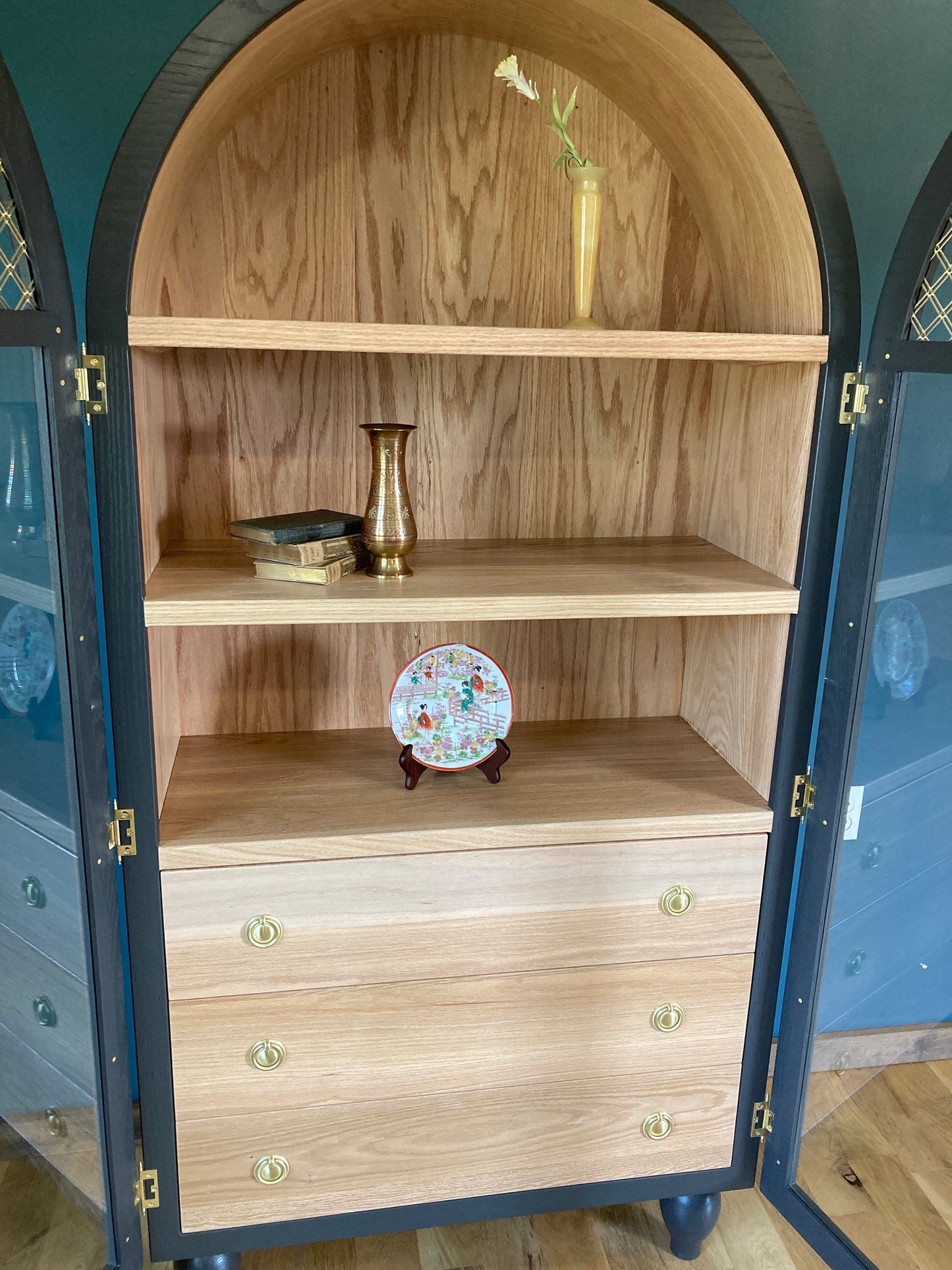 A gray cabinet with glass doors and a wood interior. The cabinet has three shelves and three drawers. The shelves are empty, but the drawers have brass handles. There is a small vase with a flower on the top shelf, a vase on the middle shelf, and a decorative plate on the bottom shelf.