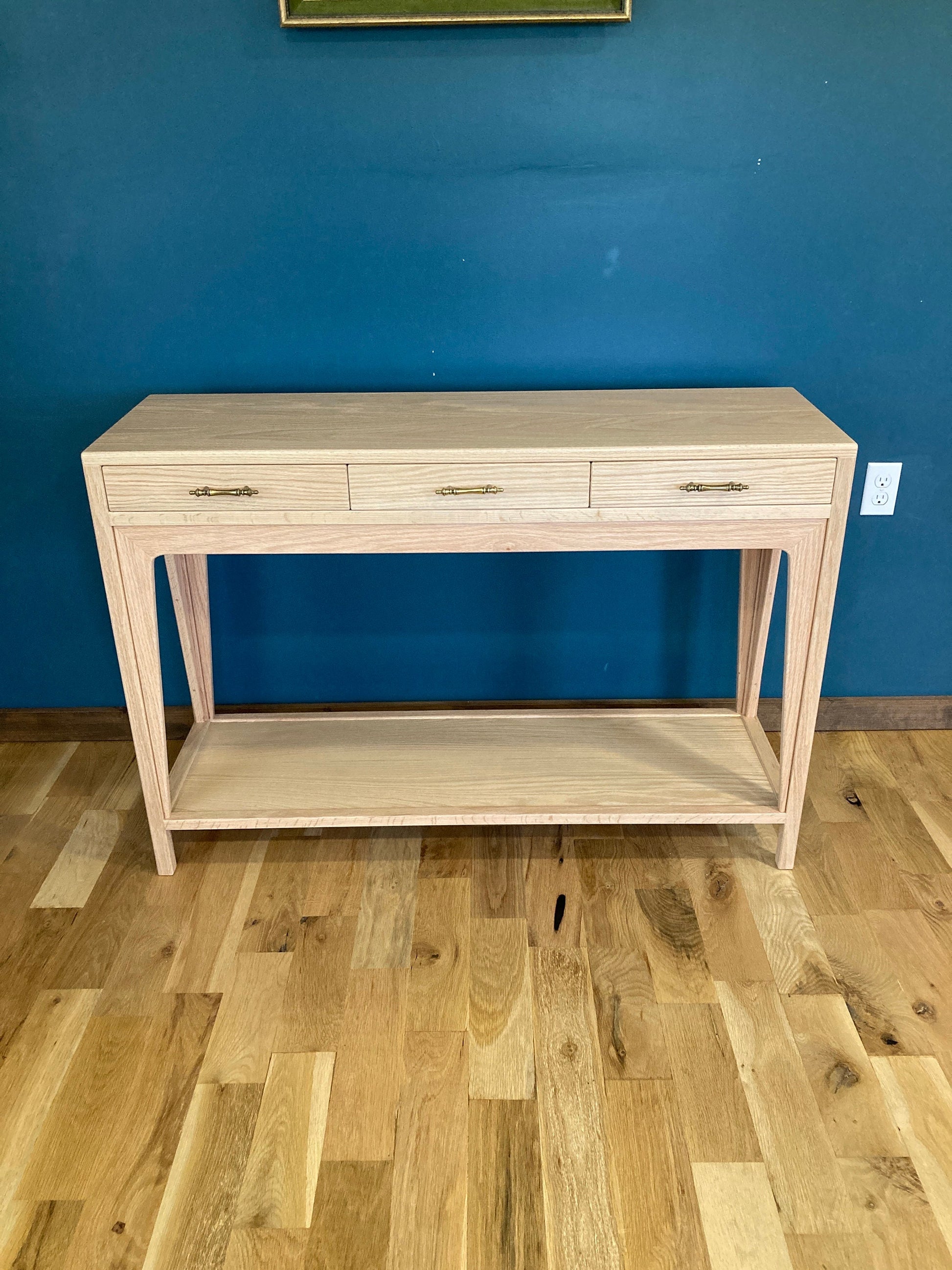 A light wood console table with three drawers and a lower shelf. The drawers have brass pulls. The table is standing on a light wood floor.