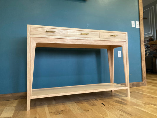 A light wood console table with three drawers and a lower shelf. The drawers have brass pulls. The table is standing on a wood floor.