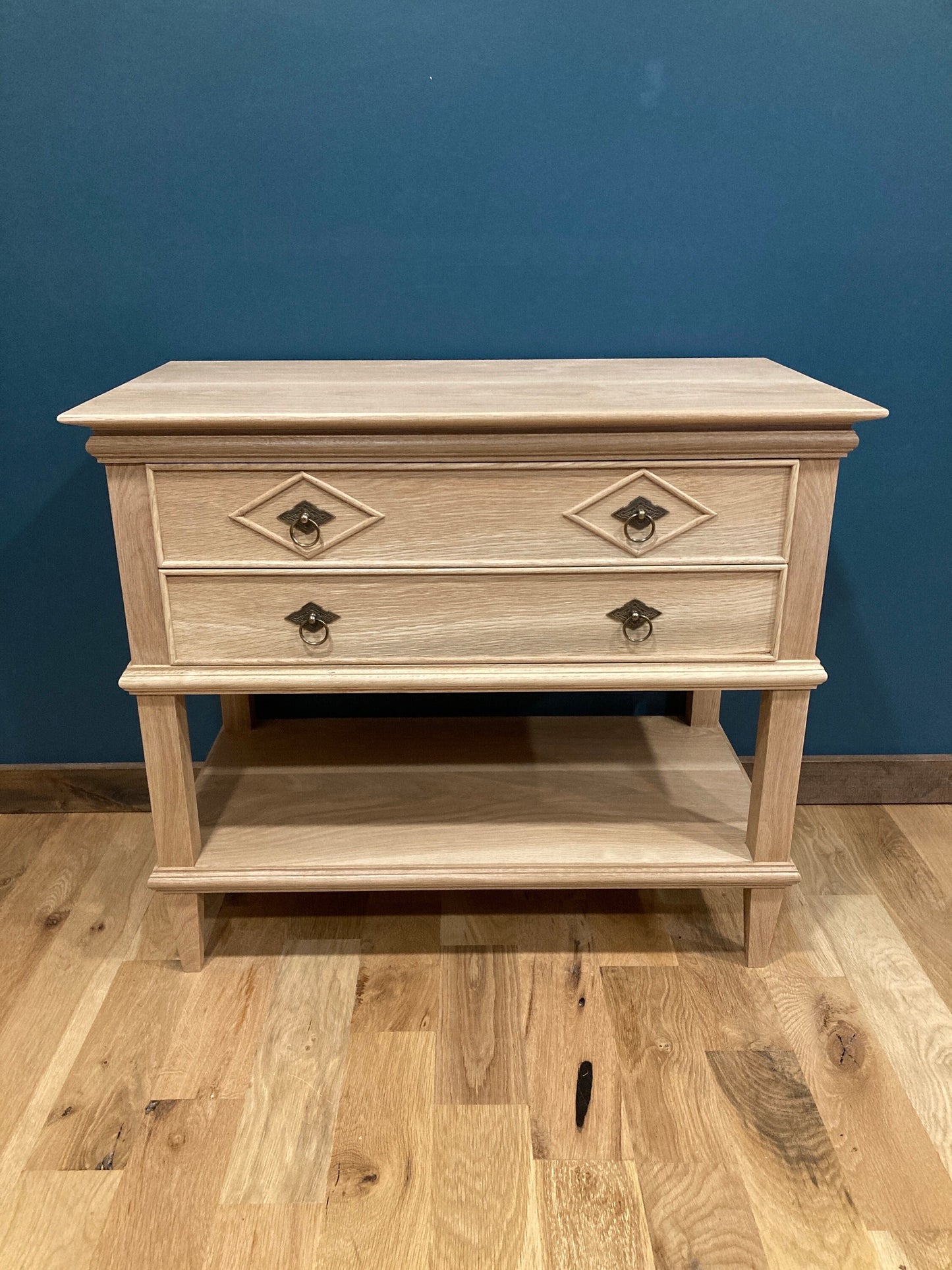 Custom hardwood nightstand with two drawers and a bottom shelf featuring diamond-patterned accents and vintage-style metal pulls, plus an open lower shelf, shown against a teal wall on hardwood flooring.