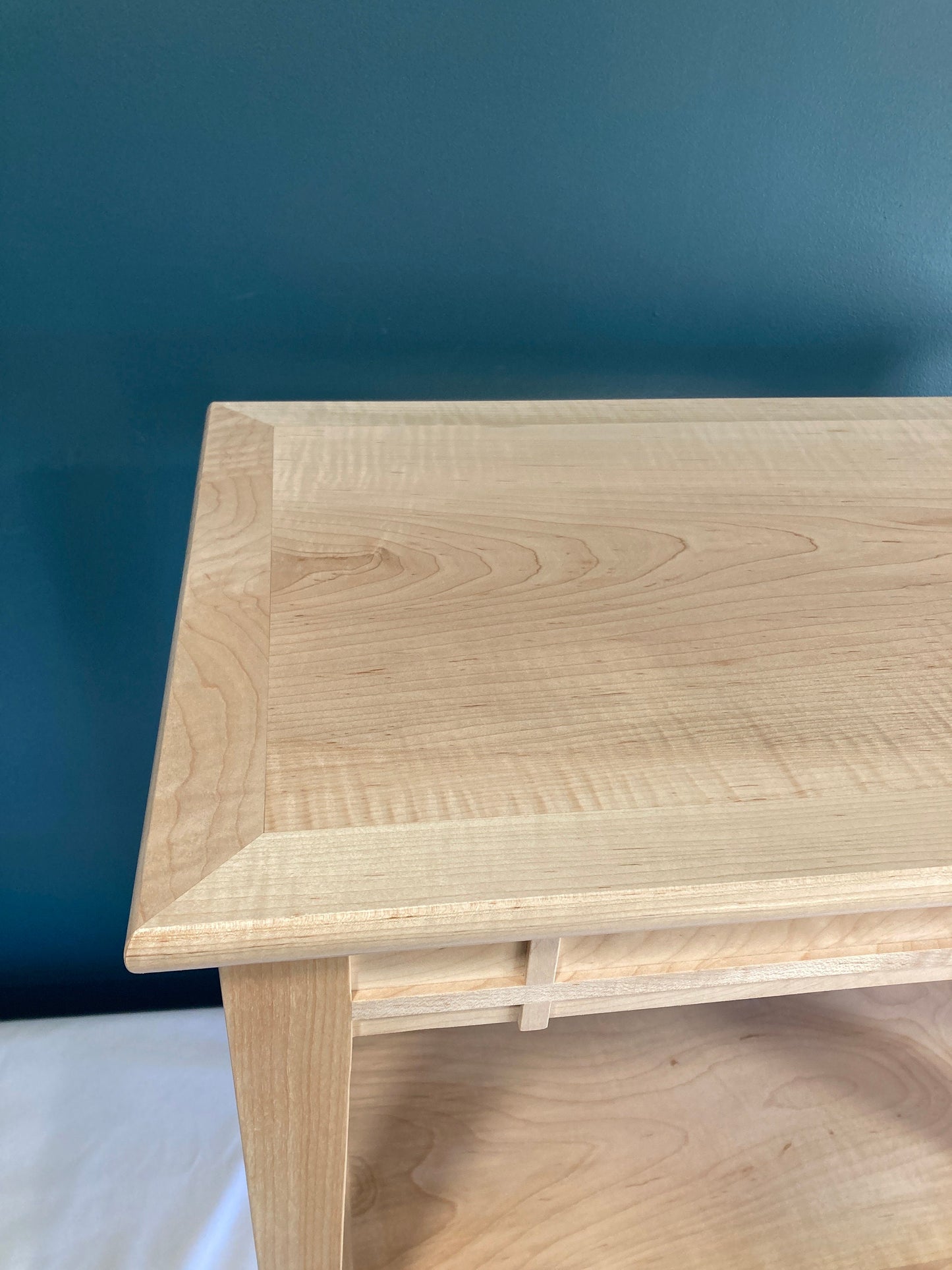 Close-up view of a light natural wood end table. The table features a smooth, light-colored wood top with a slightly raised edge.  The table's base showcases a simple, clean design with visible wood grain. The light wood is consistent throughout the piece. The table is made of solid maple, showcasing a high-quality finish. The overall style is minimalist and modern.