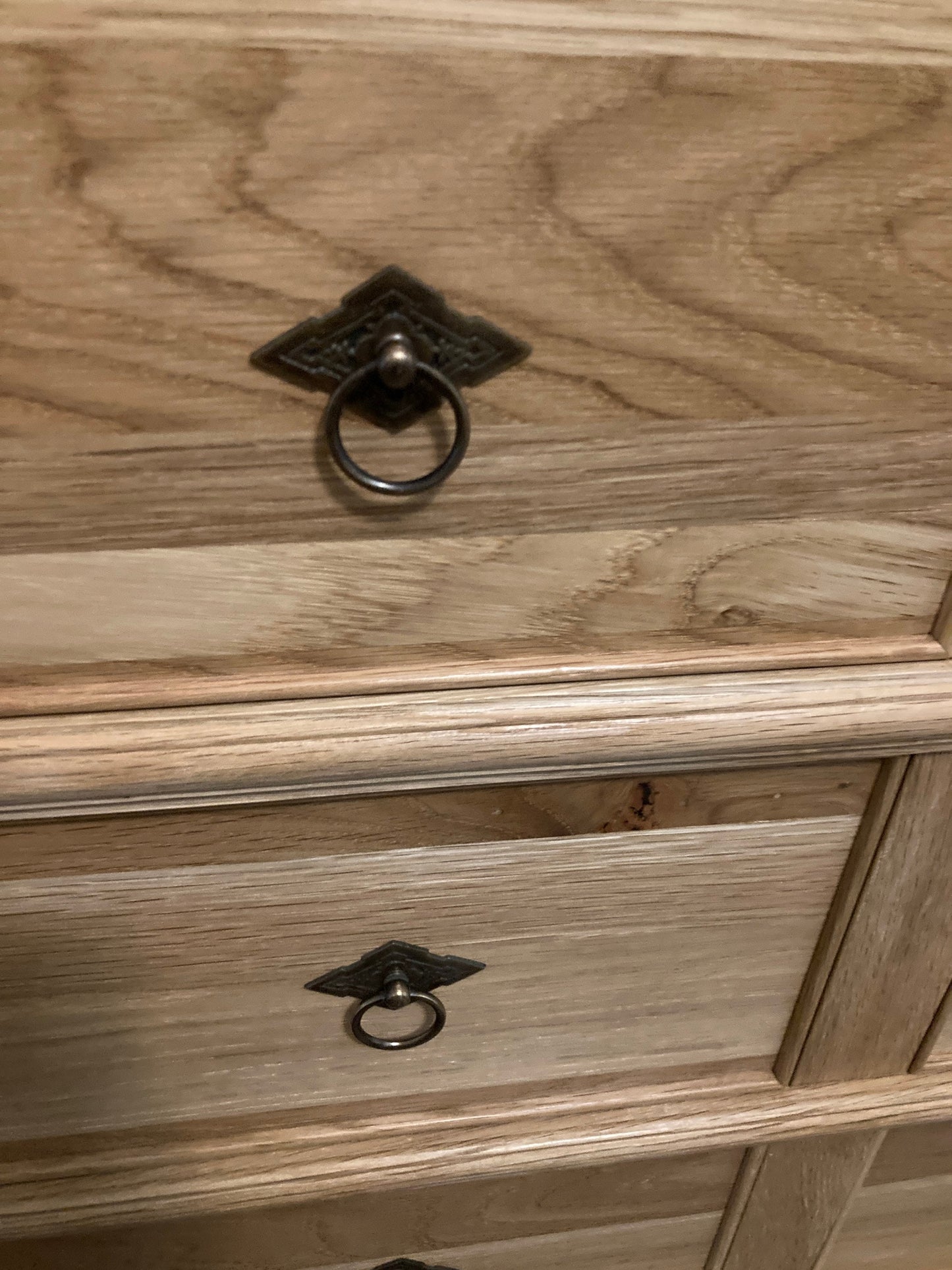 Close-up of a wooden dresser with  drawers. Each drawer has a decorative, diamond-shaped, bronze-colored handle with a ring pull. The wood grain is visible on the dresser.