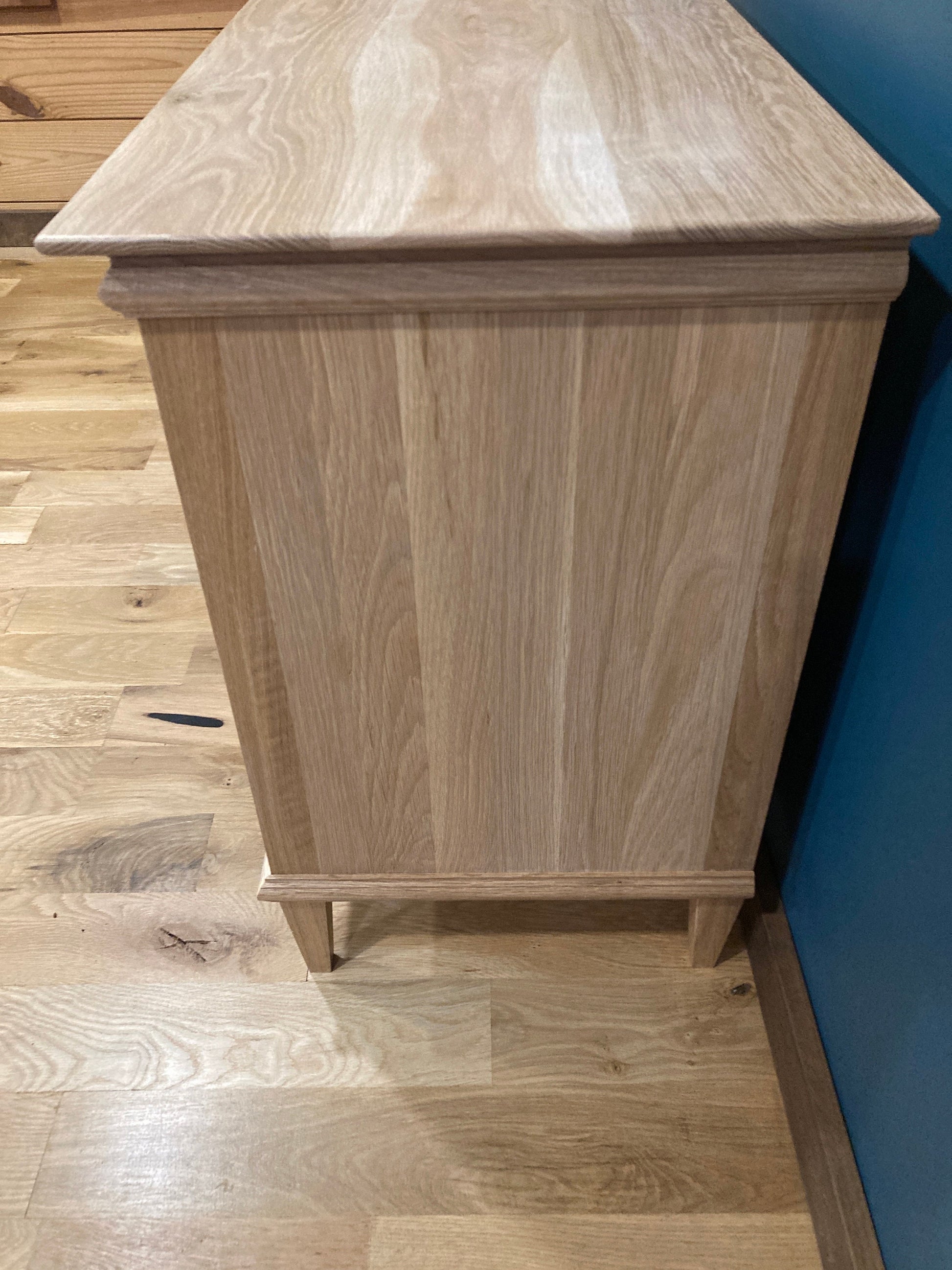 A light wood nightstand with a simple design. The nightstand has a smooth, flat top and a rectangular body with a slightly raised base. The nightstand is made of light wood with a natural grain pattern. The nightstand is standing on a light wood floor.