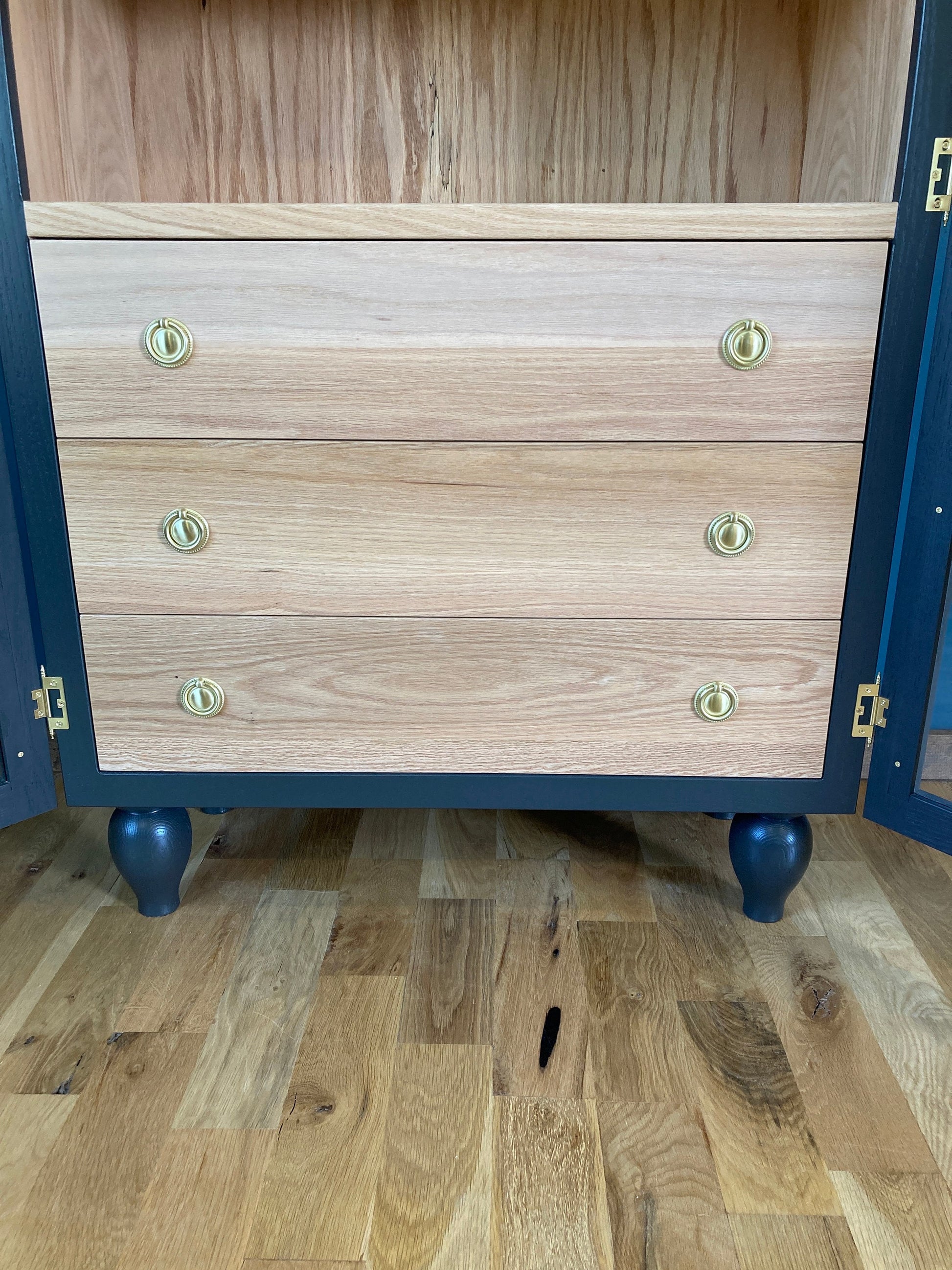 A gray cabinet with three drawers and gold knobs. The cabinet is sitting on a wooden floor. The cabinet has a wood grain finish and is made of solid wood.