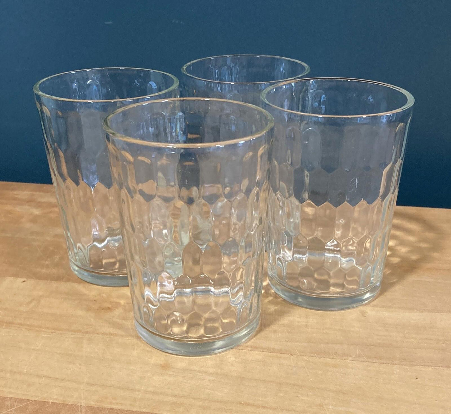 Four clear glass tumblers with textured design on a wooden surface against a blue wall.