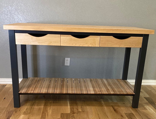 A modern kitchen island features a light wood top with three drawers and a lower shelf. The table's legs are a dark blue, creating a stylish contrast. The drawers have a subtle curved detail, adding a touch of elegance. The lower shelf is made of wood slats, providing both storage and visual interest. This versatile piece would be perfect in a kitchen.