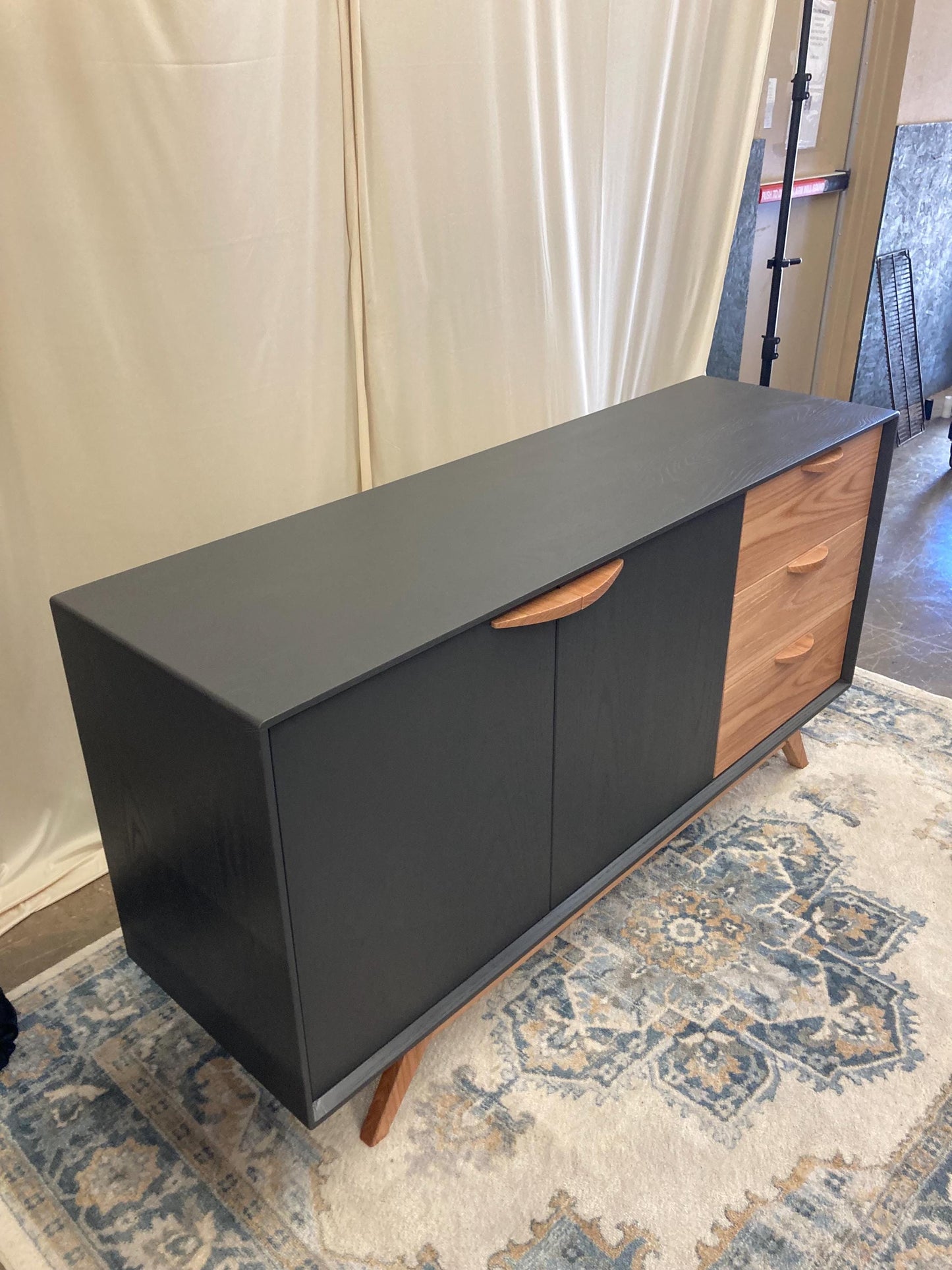 A mid-century modern sideboard with a dark gray finish and light wood accents. The sideboard features two large doors with curved wooden handles and three drawers on the right side. It sits on tapered legs and has a sleek, minimalist design. This stylish storage piece is perfect for a living room or dining room.
