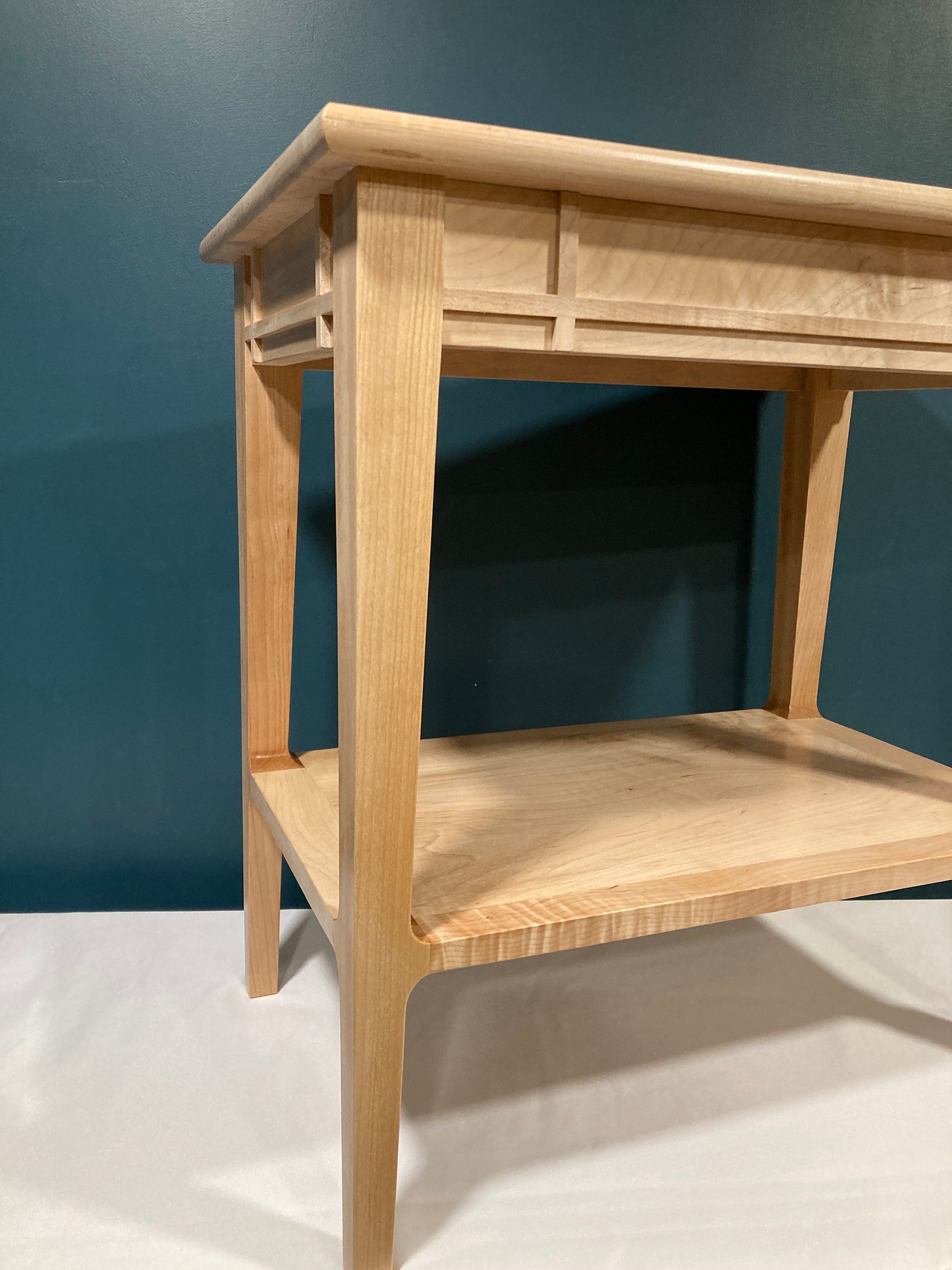 A light-colored wooden end table with a minimalist design. The table features a rectangular top with a geometric inlay, a lower shelf, and tapered legs. The wood grain is visible, showcasing the natural beauty of the material.  The piece is a simple, yet elegant addition to any room.