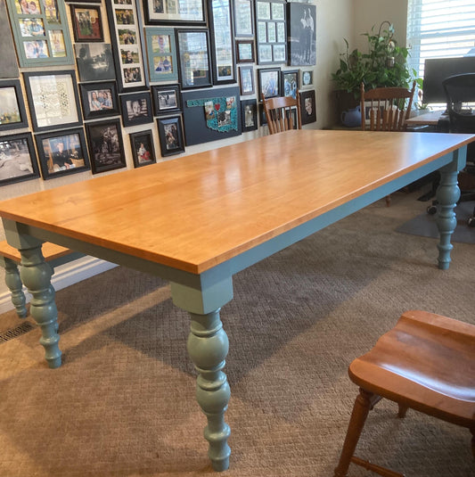 A light wood table with a blue painted base and hand turned legs. The table is in a room with a carpeted floor and a wall with framed pictures.