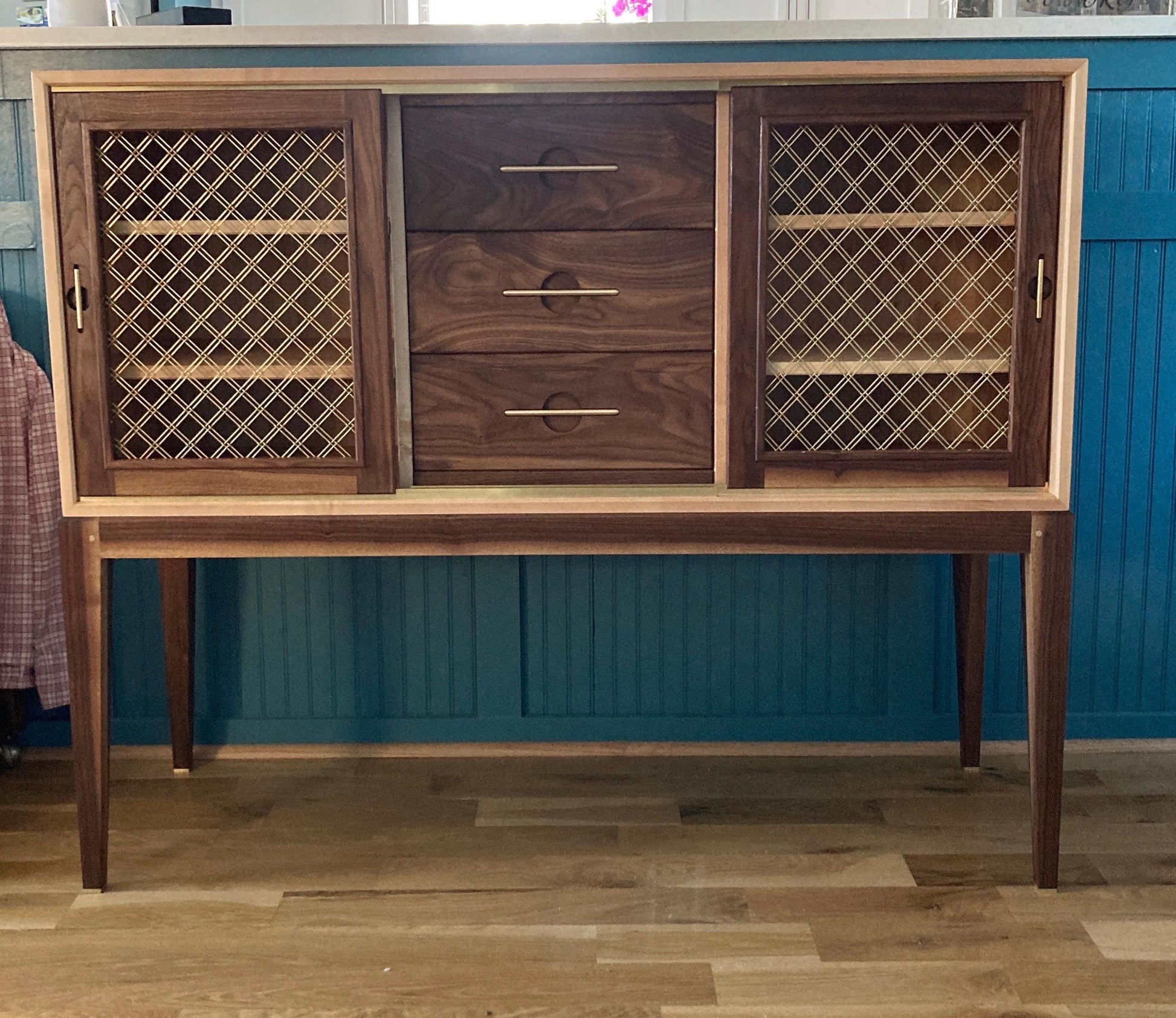  A wooden cabinet with three drawers and two doors. The doors have a brass lattice design and are made of light wood. The cabinet is sitting on four legs and has a dark wood finish. The cabinet is in front of a teal wall.