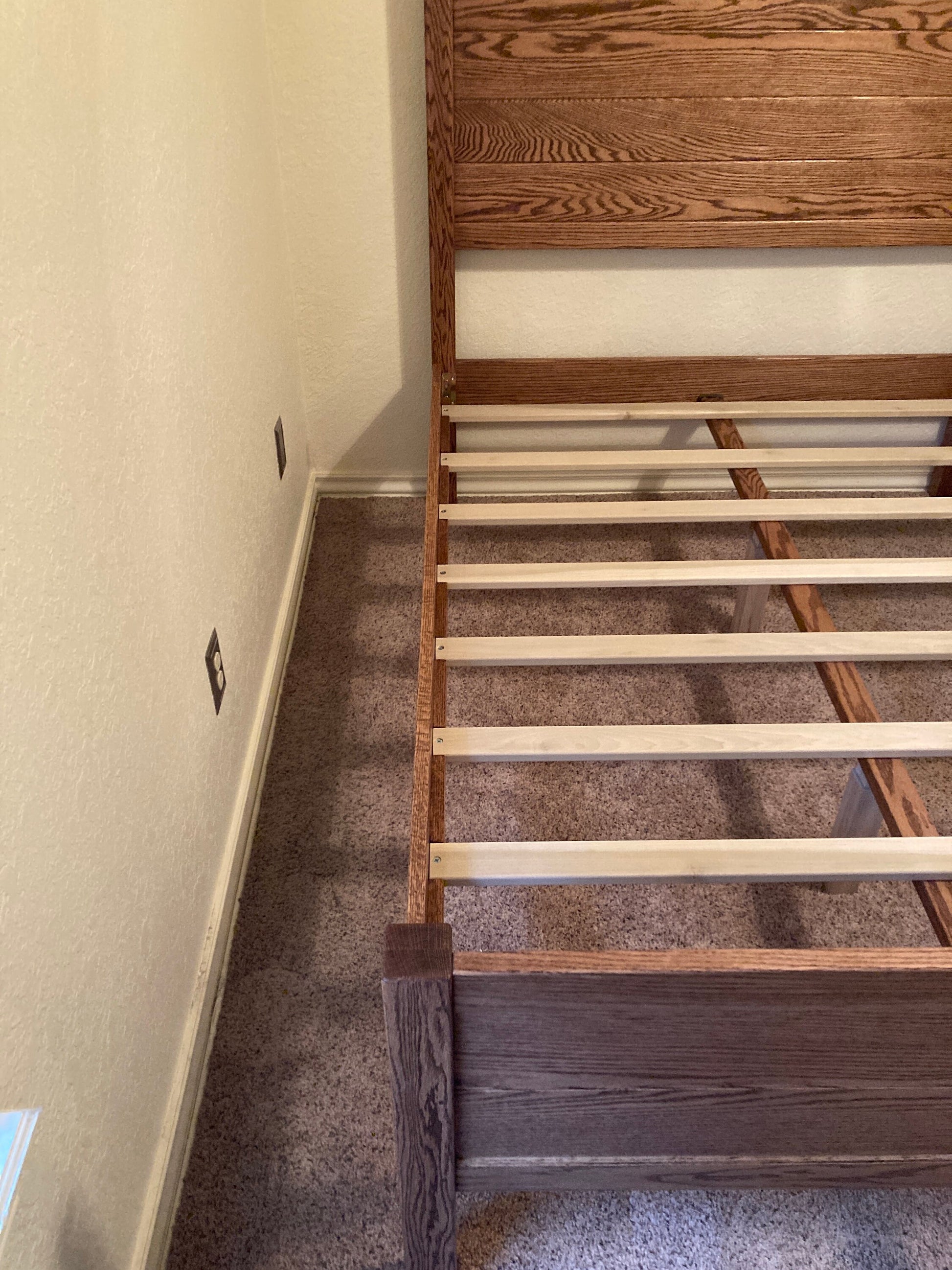 A wooden bed frame with a brown stain. The bed frame has a slatted headboard and a slatted footboard. The bed frame is sitting on a light brown carpet.
