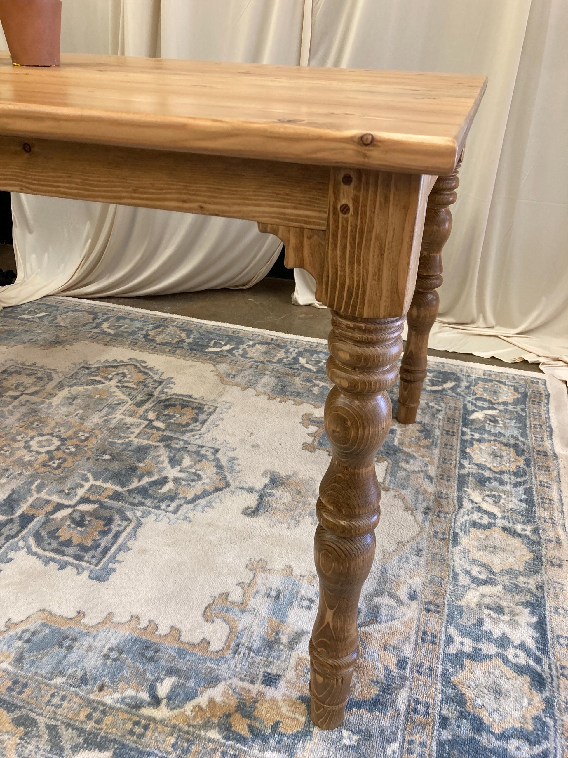 A wooden table with a light brown finish. The table has a rectangular top and four turned legs. The table is sitting on a rug with a blue and brown pattern.