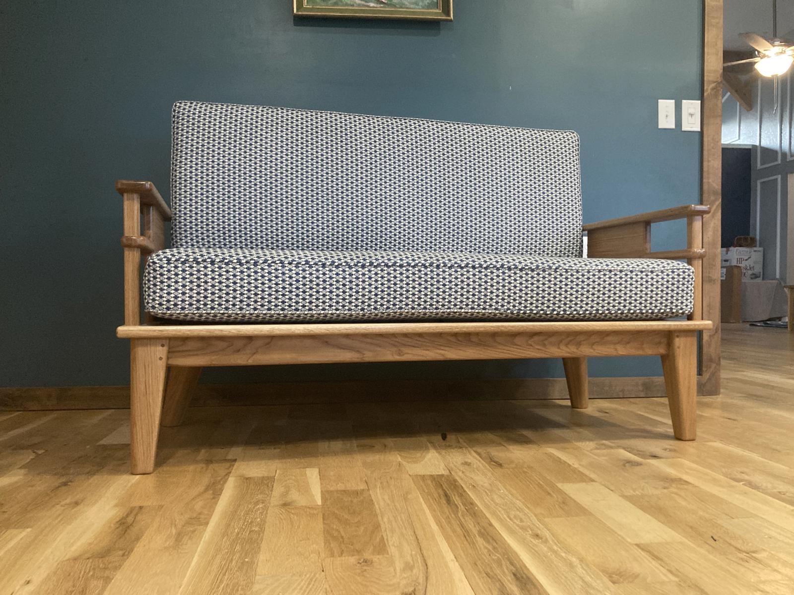 Front view of a solid wood couch with upholstered cushions.  It is sitting on a wood floor in front of a teal wall.