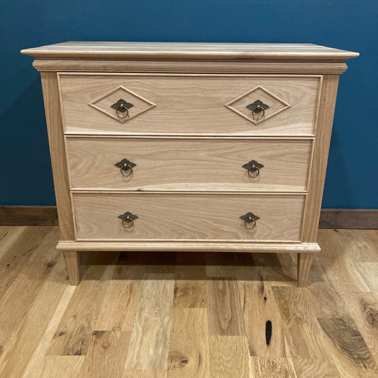 A light solid oak wood nightstand with three drawers and ornate metal handles. The dresser is in front of a blue wall and a wood floor.