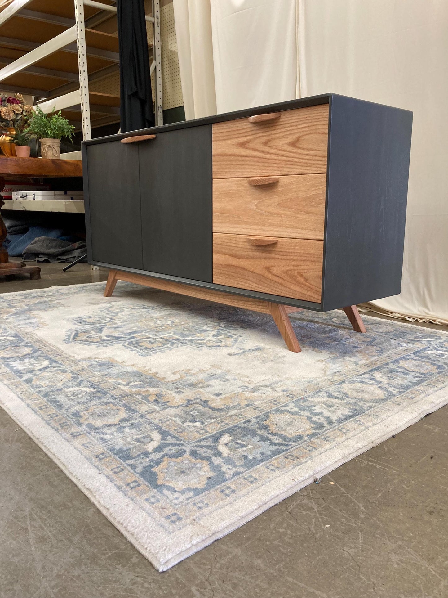 A mid-century modern sideboard with a two-toned design. The left side features dark gray painted wood doors, while the right side showcases three drawers in light oak wood.  The sideboard has tapered legs and simple, understated wood handles. This stylish storage solution is perfect for a living room or dining room.