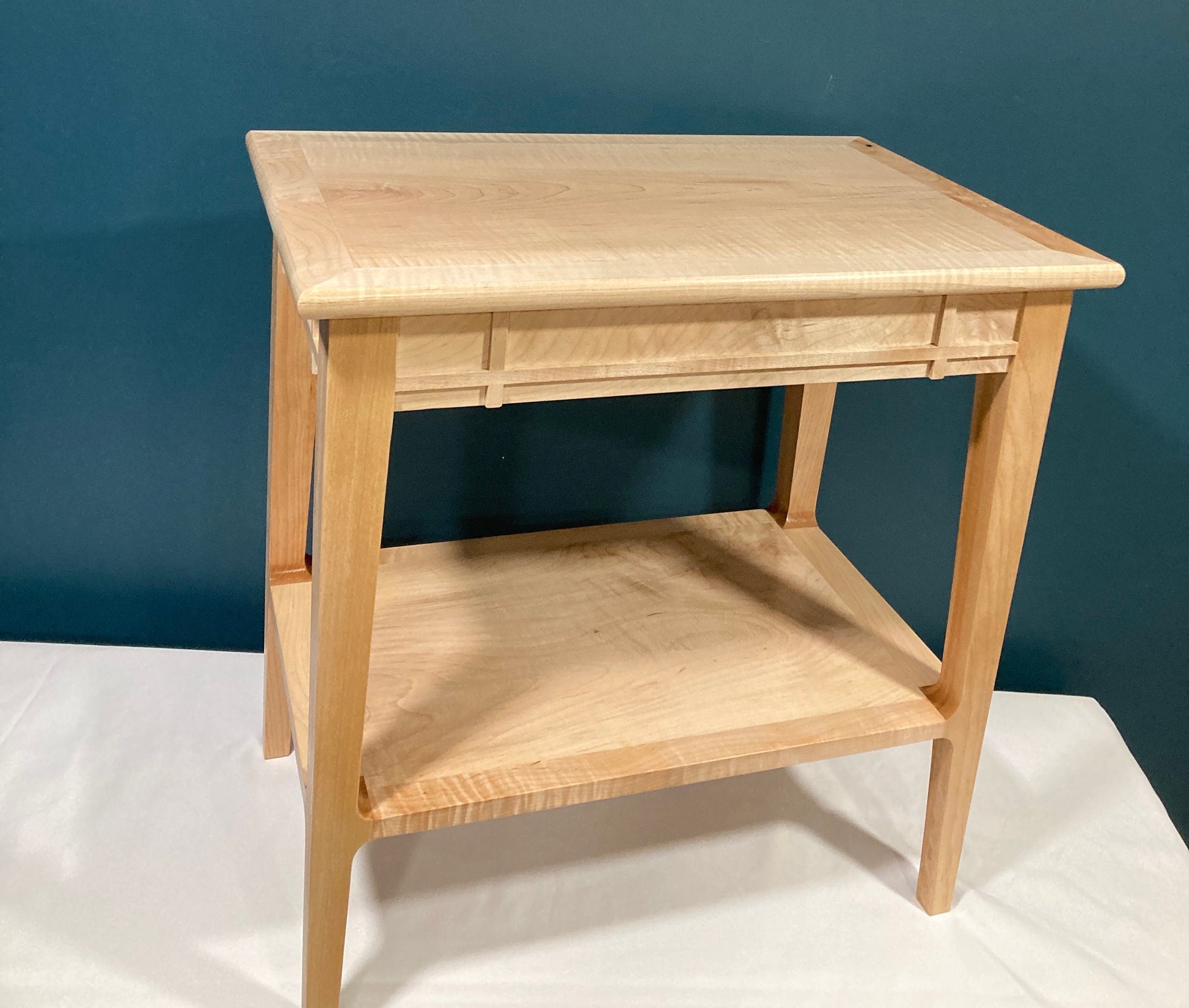 A light-colored wooden end table with a minimalist design. The table features a rectangular top and a lower shelf, both made of light-colored wood with a subtle grain pattern.  The table's legs are straight and slender, and the top has a simple, clean edge. The overall style is modern and functional, suitable for a living room or bedroom. Made of solid maple wood. 