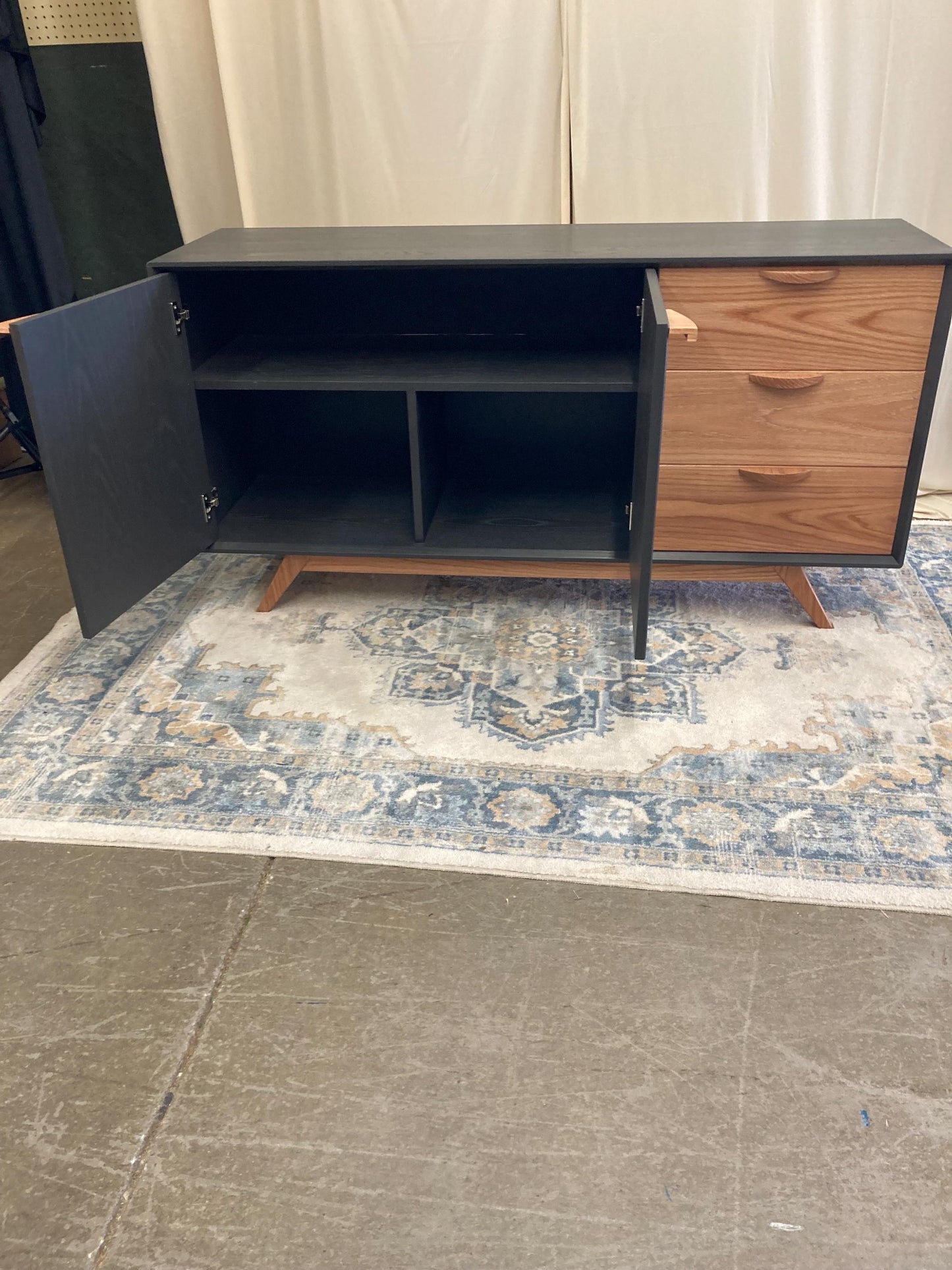 A mid-century modern sideboard with a dark gray finish and light wood drawers. The cabinet features two doors that open to reveal interior shelving, and three drawers on the right side.  The sideboard sits on tapered legs and is displayed on a patterned rug. The piece is a stylish storage solution for living rooms or dining rooms.