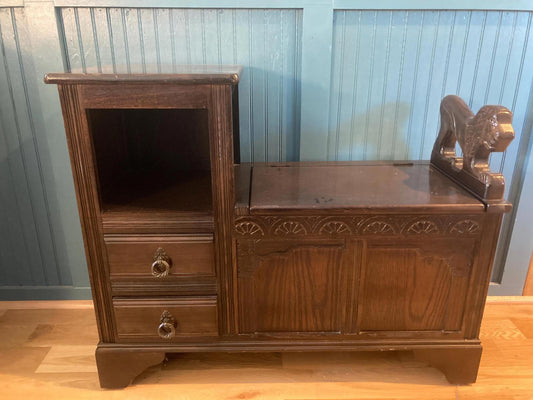 A dark brown wooden telephone table with a carved lion head detail. The table features a cabinet with two drawers, an open shelf, and a hinged top.  The piece is an antique, and shows some signs of age and wear. The wood grain is visible, and the overall style is traditional and ornate.