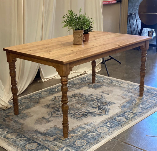 A light brown wooden table with turned legs. The table is on a rug with a blue, beige, and white pattern. There are two potted plants on the table.